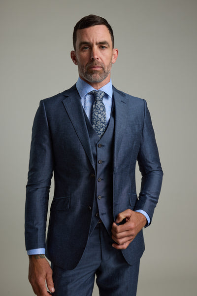 A man with short dark hair and a beard stands against a plain background, confidently wearing Barkers' Charles Texture Waistcoat with an adjustable back buckle, paired with a blue three-piece suit, light blue dress shirt, and patterned tie.