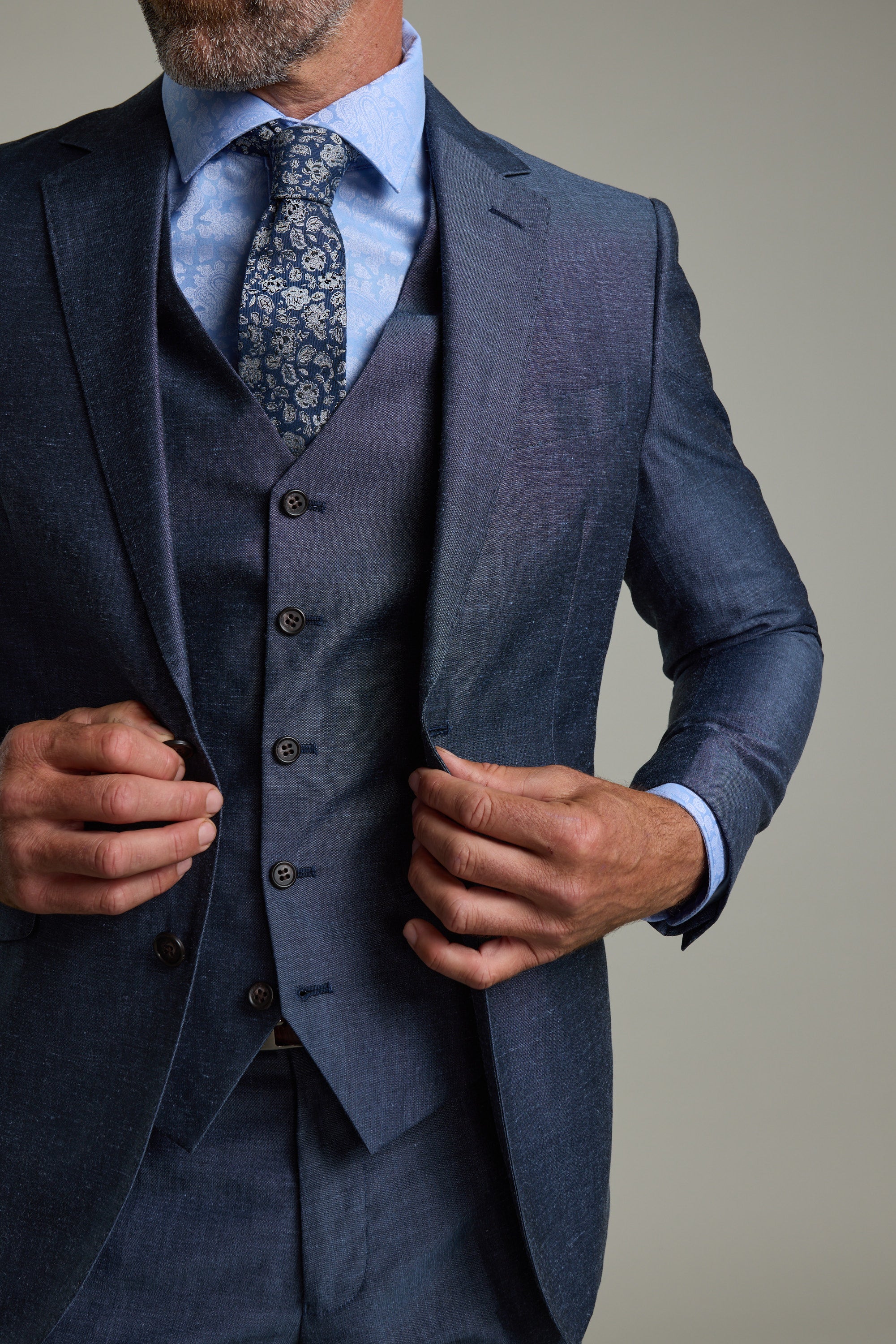 A man in a tailored dark blue three-piece suit, featuring the Barkers Charles Texture Waistcoat, wears a light blue shirt and patterned tie as he adjusts his jacket. The image is cropped at the neck and knees.