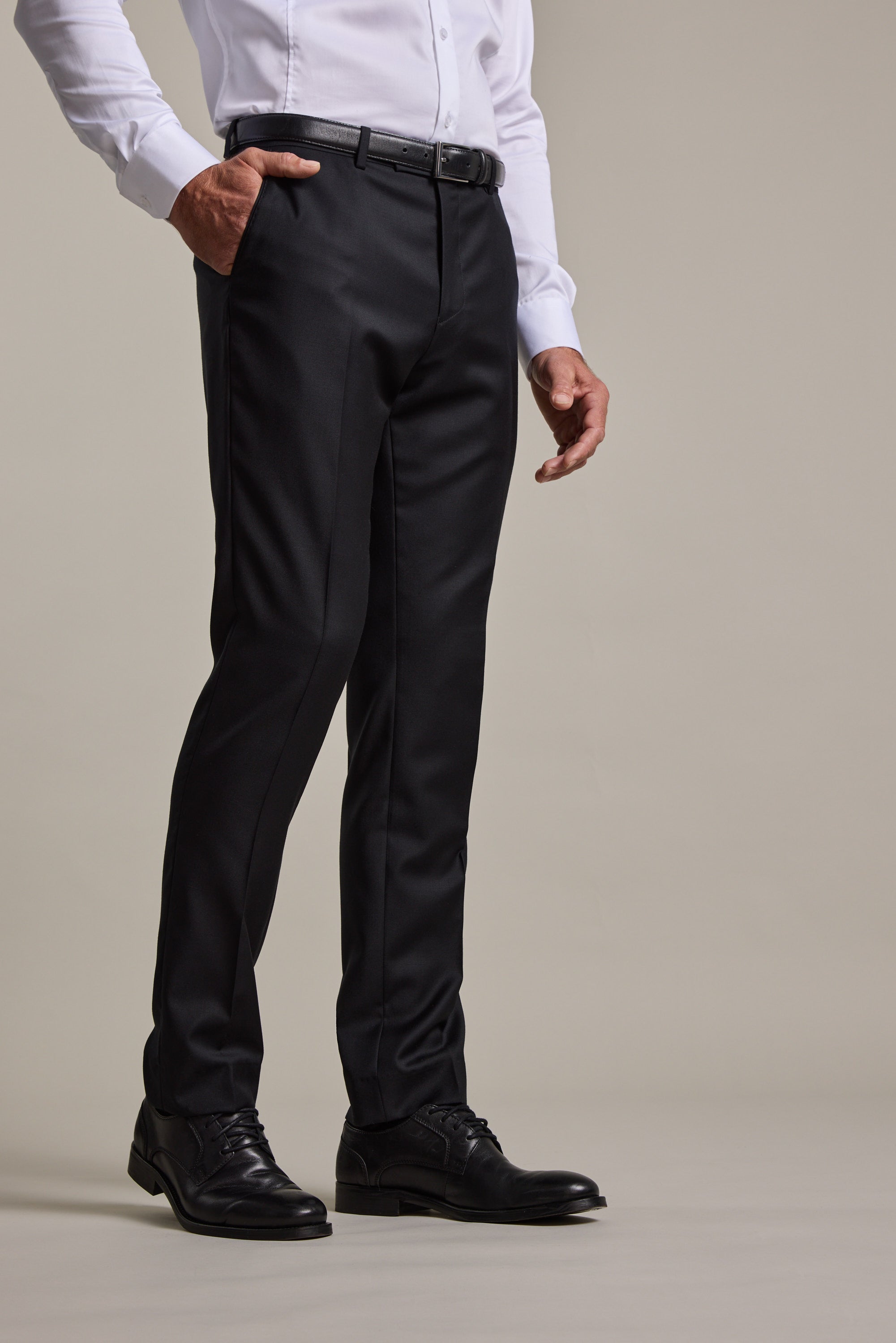 A man stands with one hand in his pocket against a plain light background, wearing Barkers' Mayfair ZQrx Suit Trouser paired with a white dress shirt, black belt, and black dress shoes.