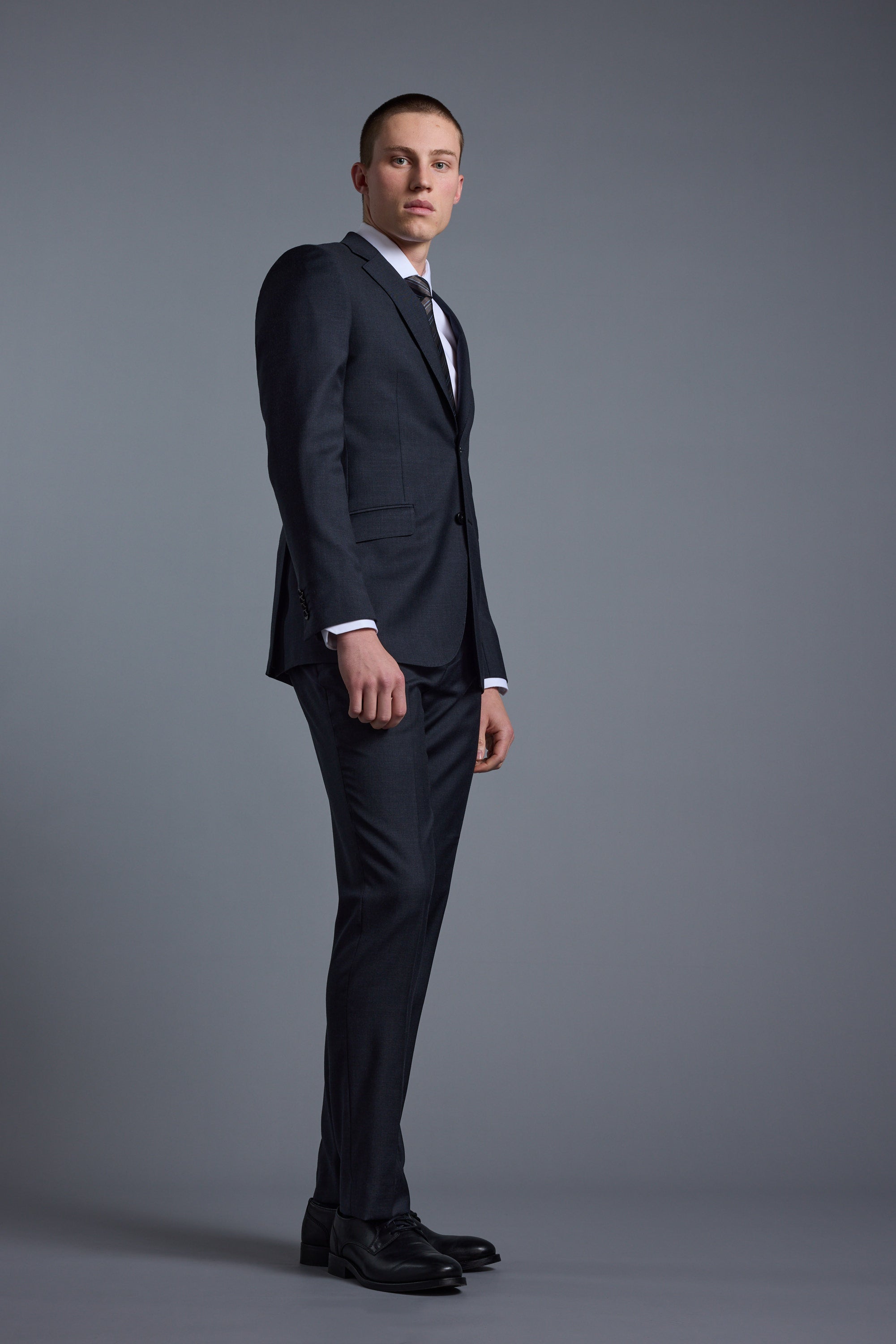 A young man in Barkers' Mayfair ZQ Suit Trouser, paired with a dark three-piece suit, white dress shirt, and tie, stands against a plain gray background, gazing slightly left with a serious expression.