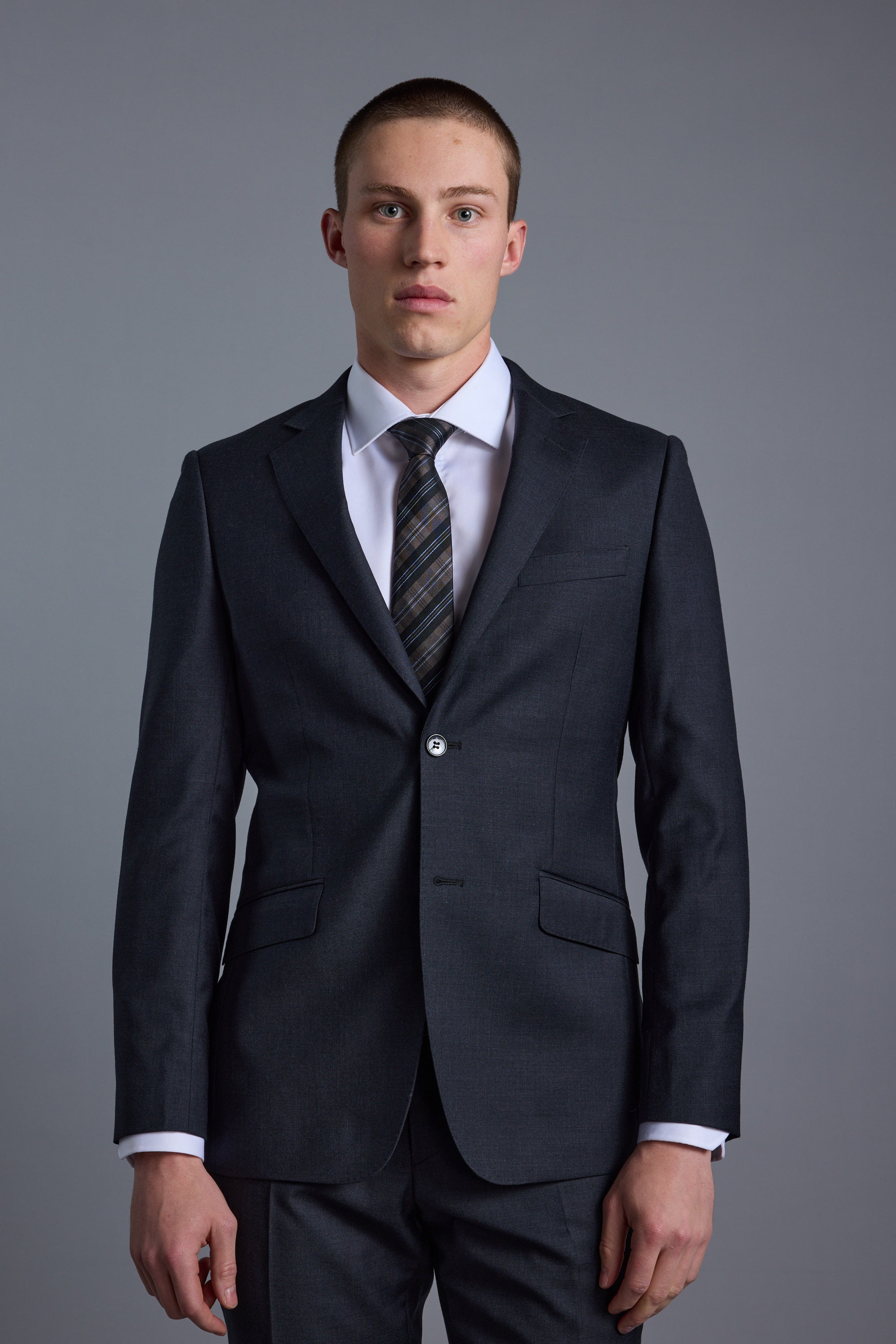 A young man with short hair and a neutral expression stands facing forward against a plain gray background, wearing the Barkers Mayfair ZQ Suit Jacket made from ZQ Merino Wool.