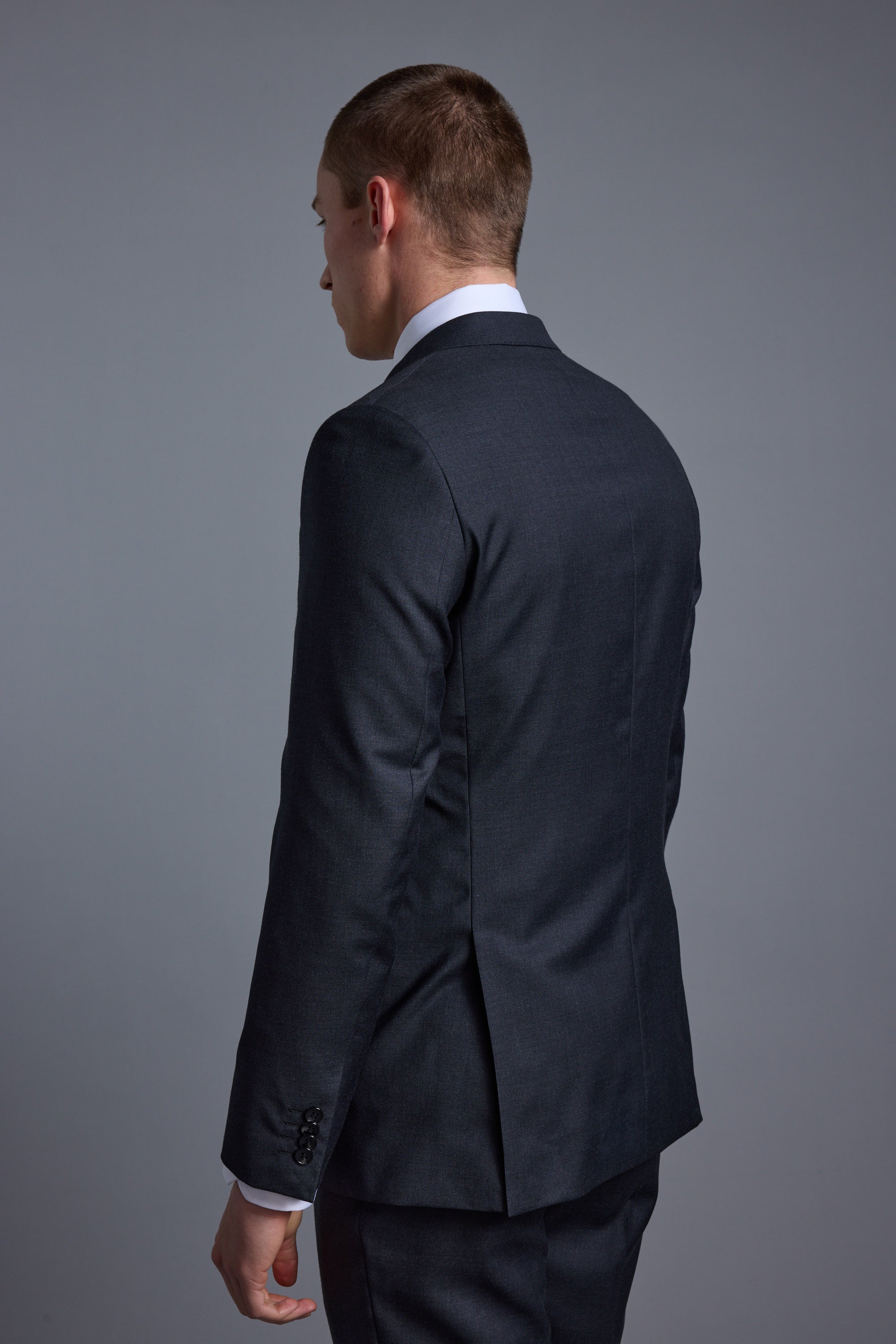A man in a dark Barkers Mayfair ZQ Suit Jacket made from breathable wool stands with his back to the camera, head slightly turned left, set against a plain gray background.