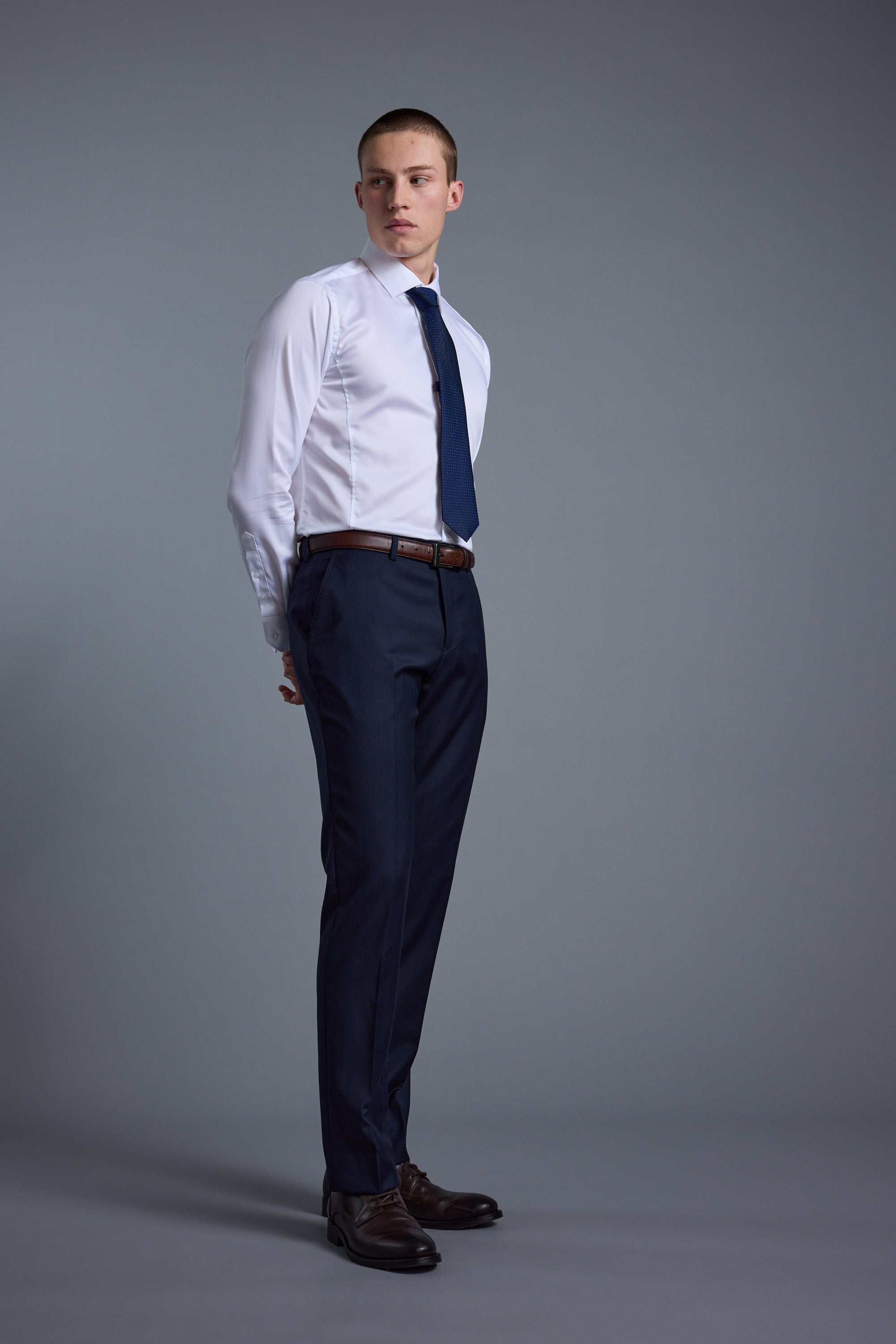 A young man stands against a gray background, wearing Barkers’ Mayfair ZQ Suit Trouser, a white dress shirt, navy blue tie, brown belt, and brown shoes. He looks to the side with his hands behind his back.