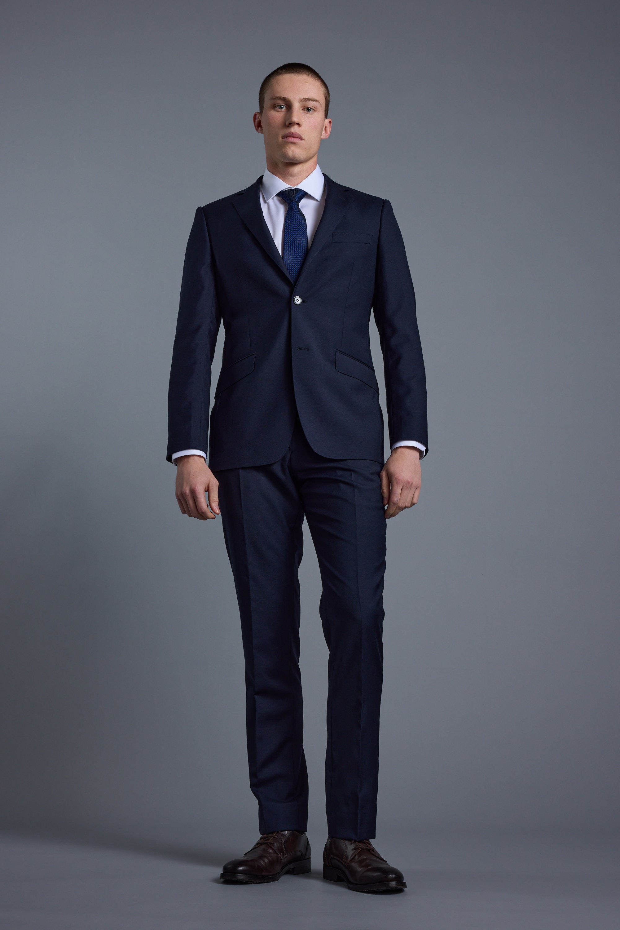 A man stands against a plain grey background wearing Barkers' Mayfair ZQ Suit Trouser in navy, made from New Zealand merino wool, with tailored fit and neutral expression, his arms resting at his sides.