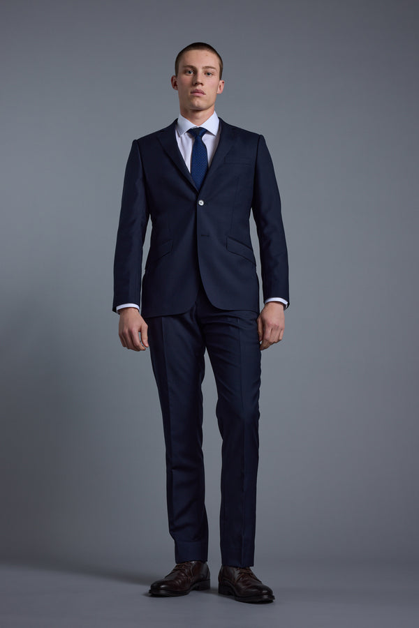 A man stands against a plain grey background wearing Barkers' Mayfair ZQ Suit Trouser in navy, made from New Zealand merino wool, with tailored fit and neutral expression, his arms resting at his sides.