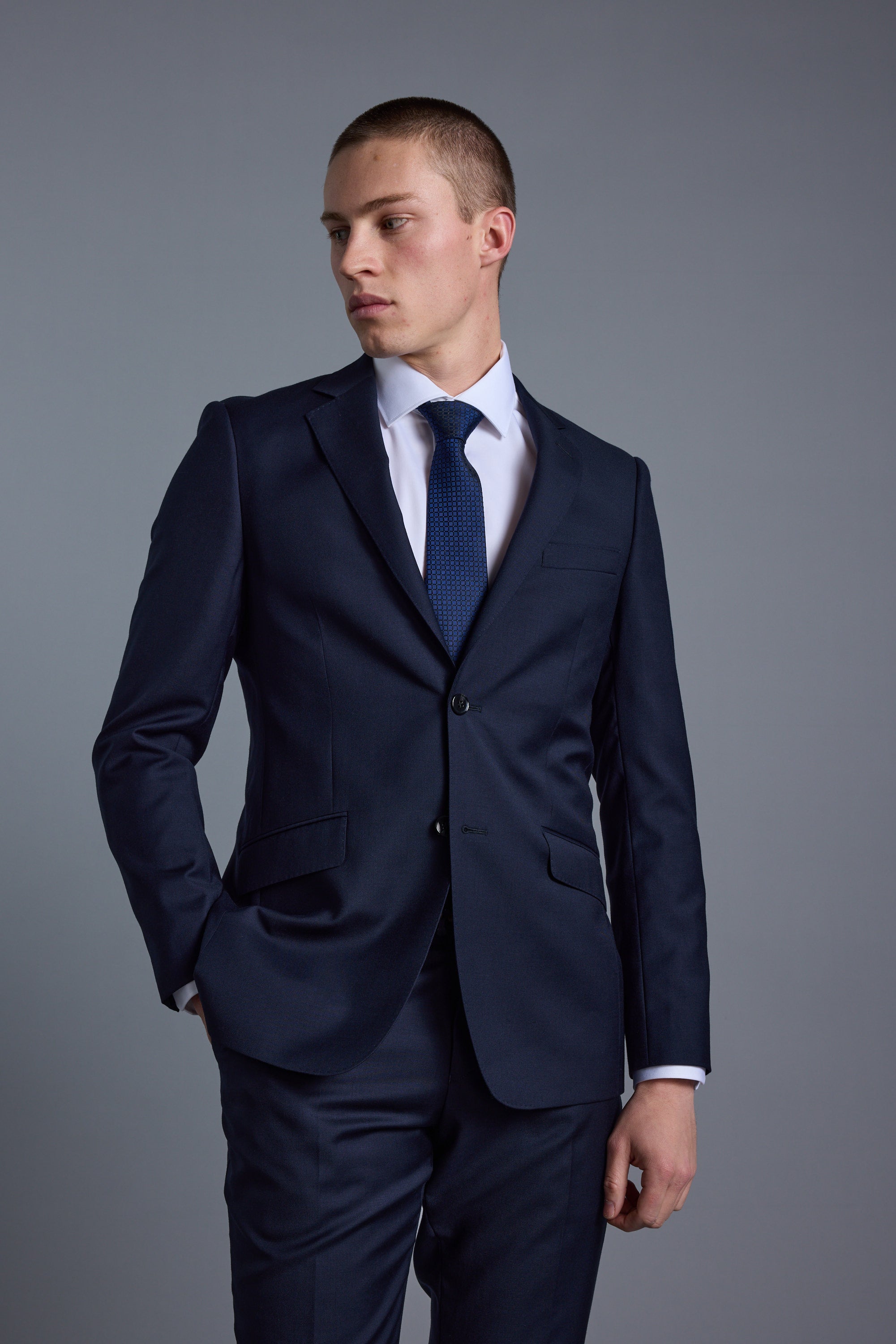 A young man wearing the Barkers Mayfair ZQ Suit Jacket, a white dress shirt, and a blue tie stands against a plain gray background, looking to his left with one hand in his pocket.