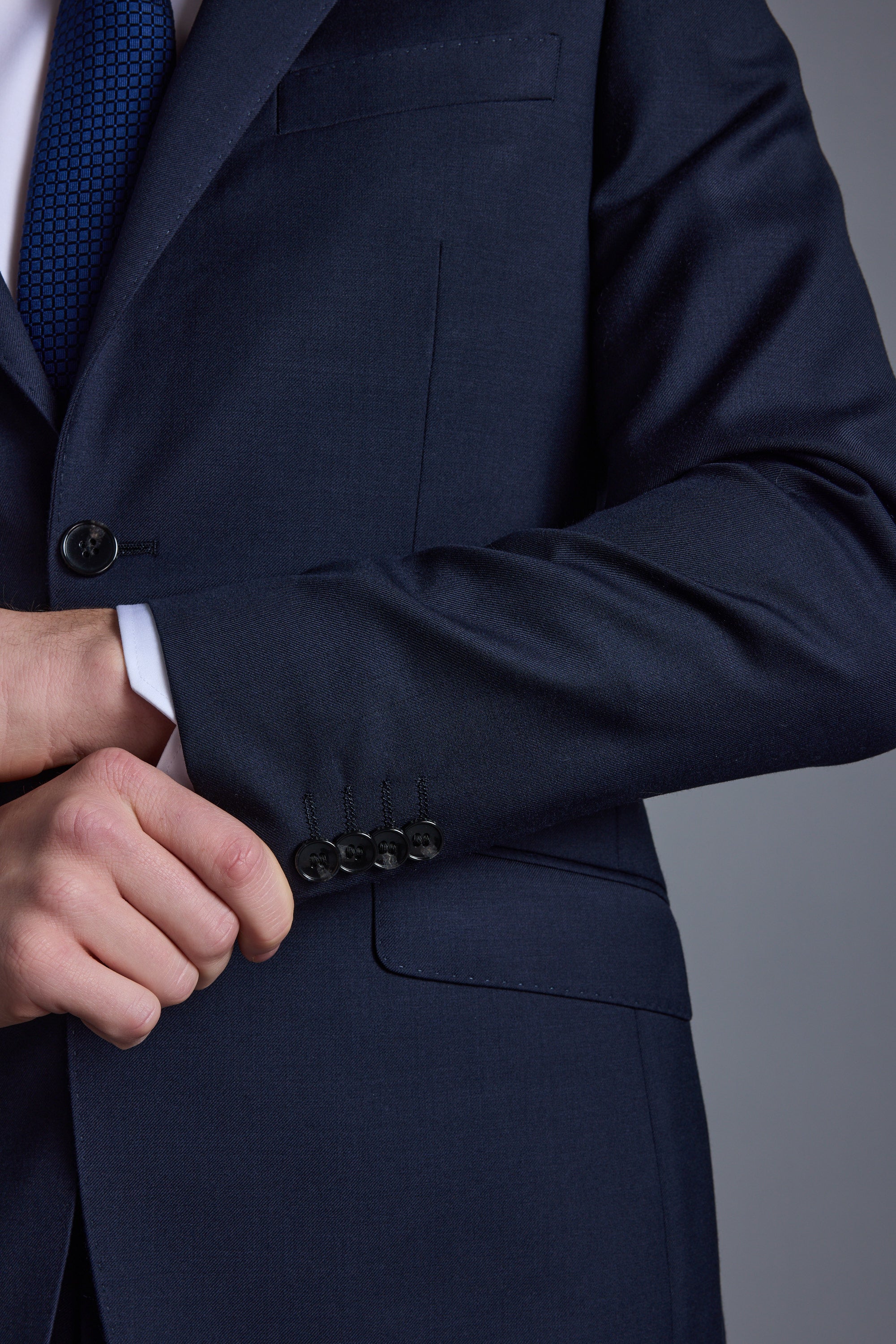 A person in a Barkers Mayfair ZQ Suit Jacket and blue patterned tie adjusts their jacket sleeve, focusing on the hand and sleeve buttons.