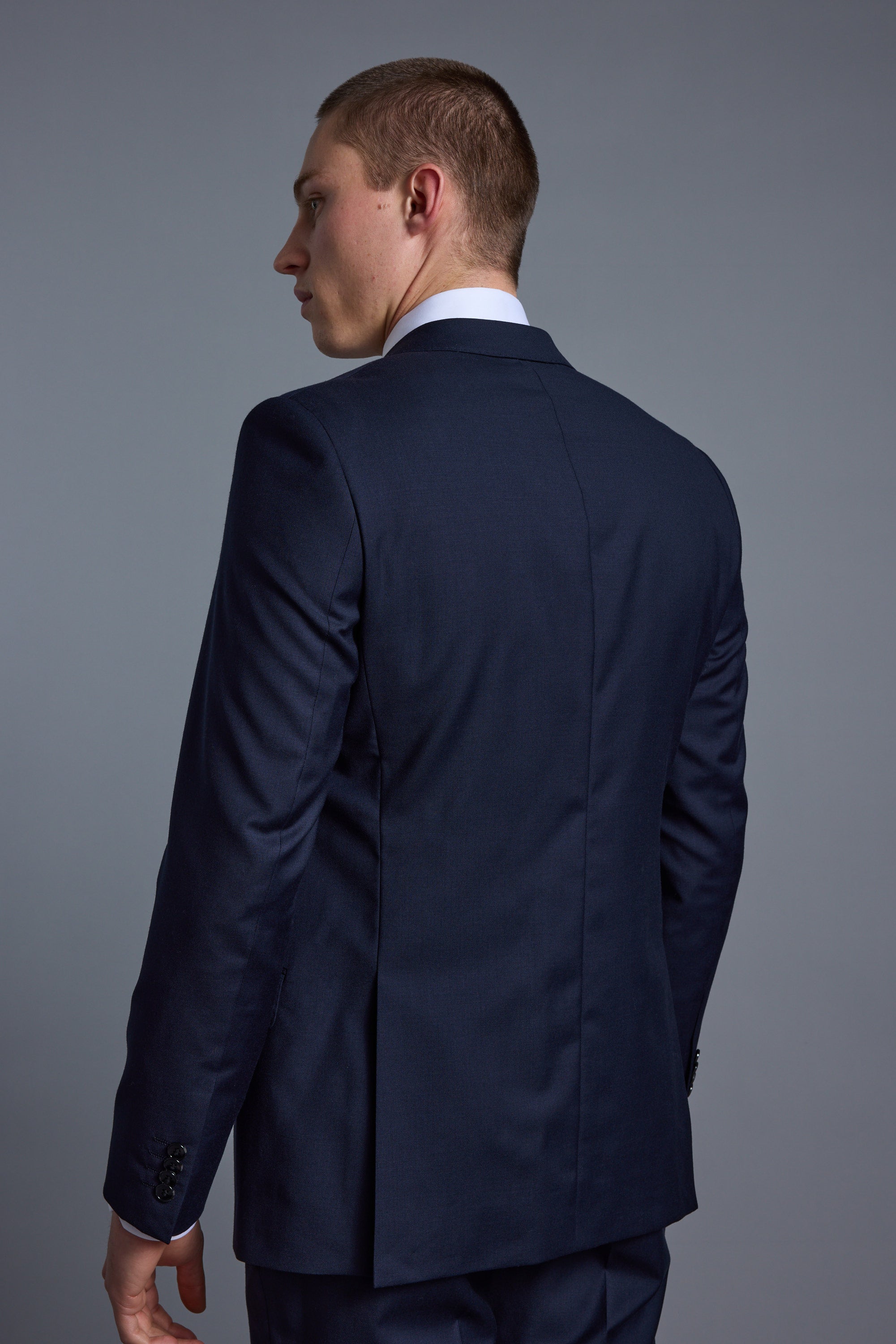 A man wearing a Barkers Mayfair ZQ Suit Jacket in dark blue stands with his back to the camera, head turned slightly to the side, against a plain gray background.