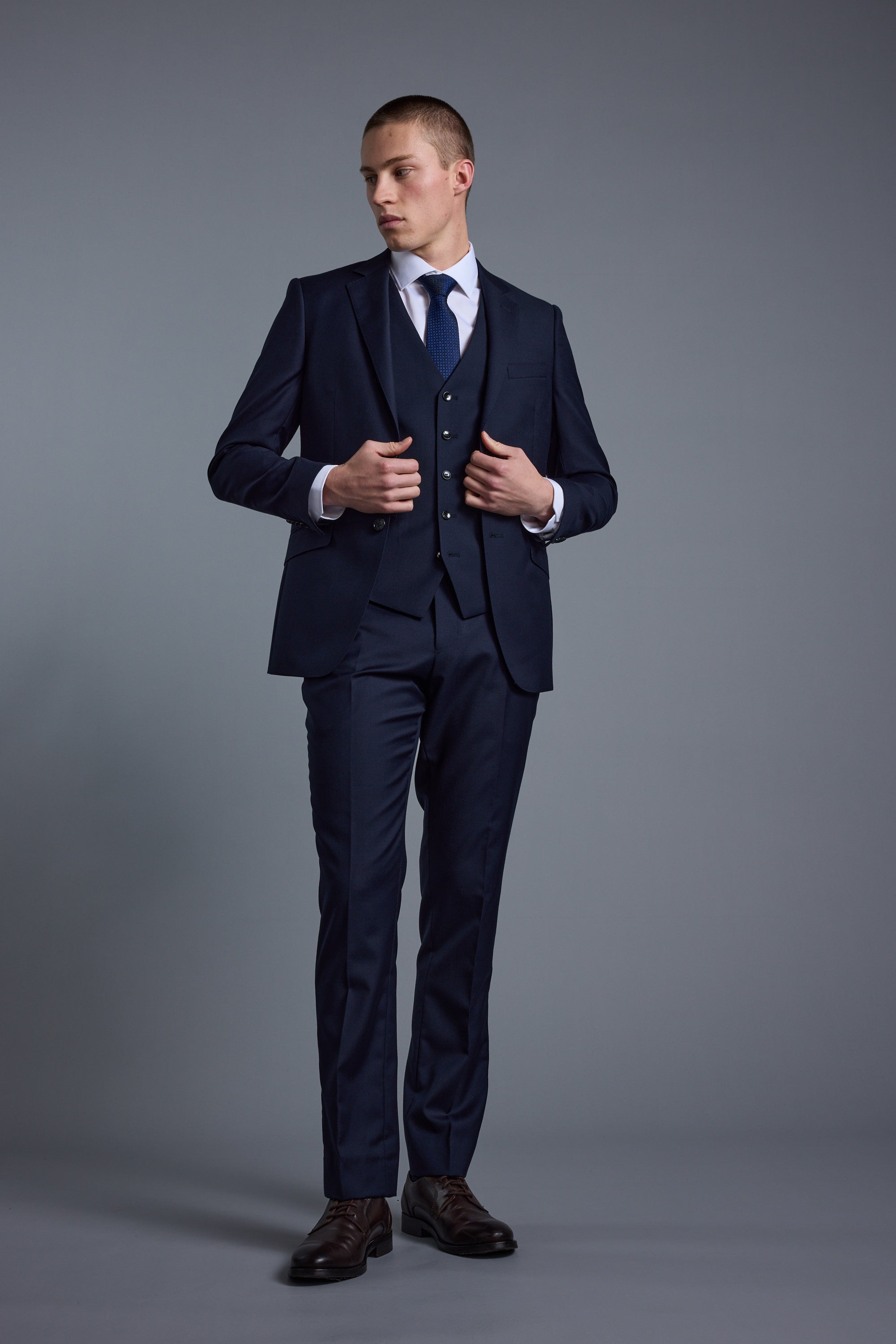 A man in a dark blue Barkers Mayfair ZQ Suit Jacket, light blue shirt, and tie stands against a plain gray background, looking to his left while adjusting his vest.