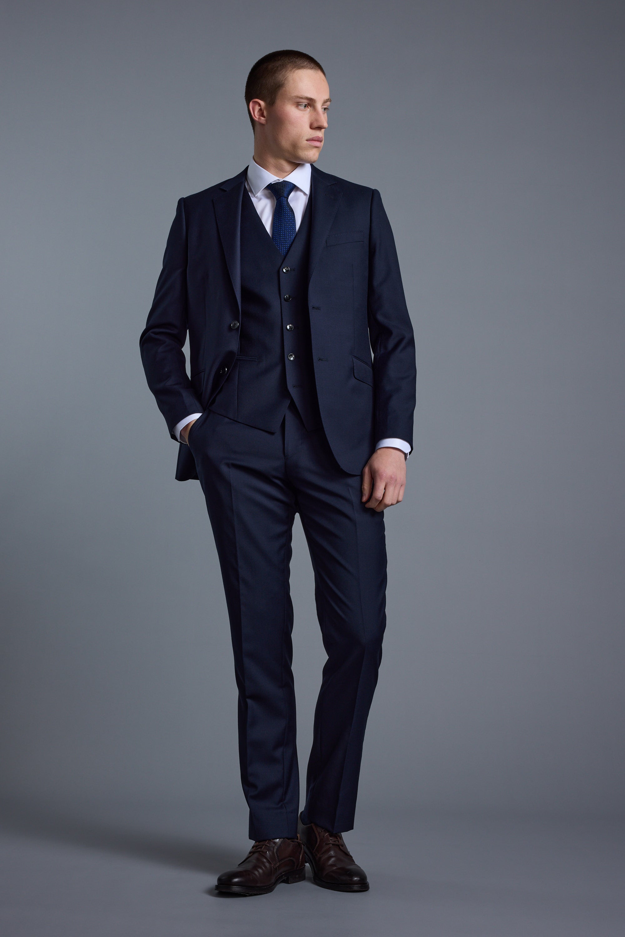 A man stands in a studio wearing Barkers’ Mayfair ZQ Suit Trouser, part of a dark blue three-piece suit made from ZQ-certified New Zealand merino wool, styled with a white shirt, blue tie, and brown dress shoes against a plain gray background.