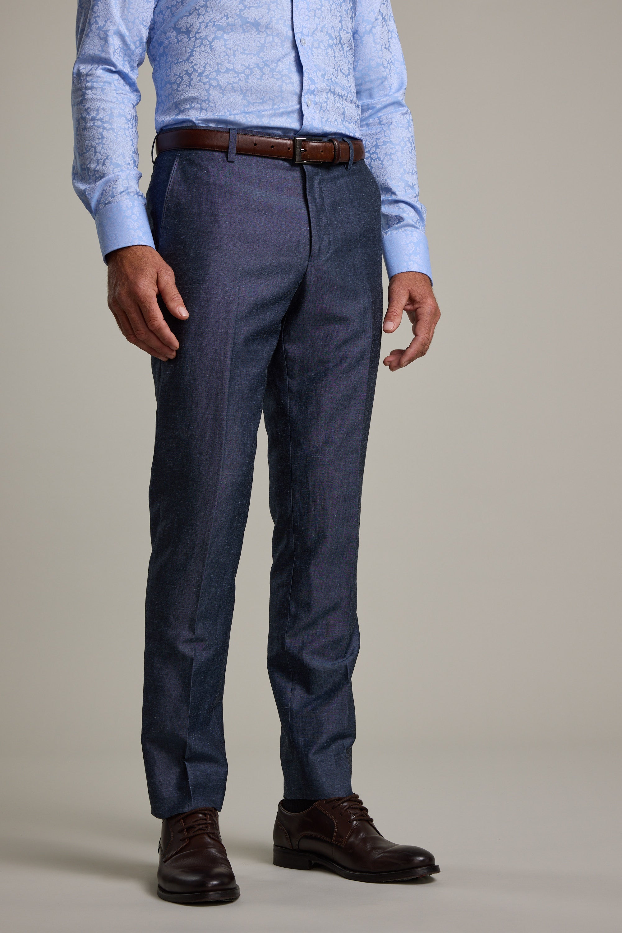 A man wears Barkers Charles Texture Suit Trouser in dark blue, a light blue patterned shirt, brown belt, and dark brown dress shoes—styled for a modern look—standing against a plain light background. Image is cropped at shoulder height.