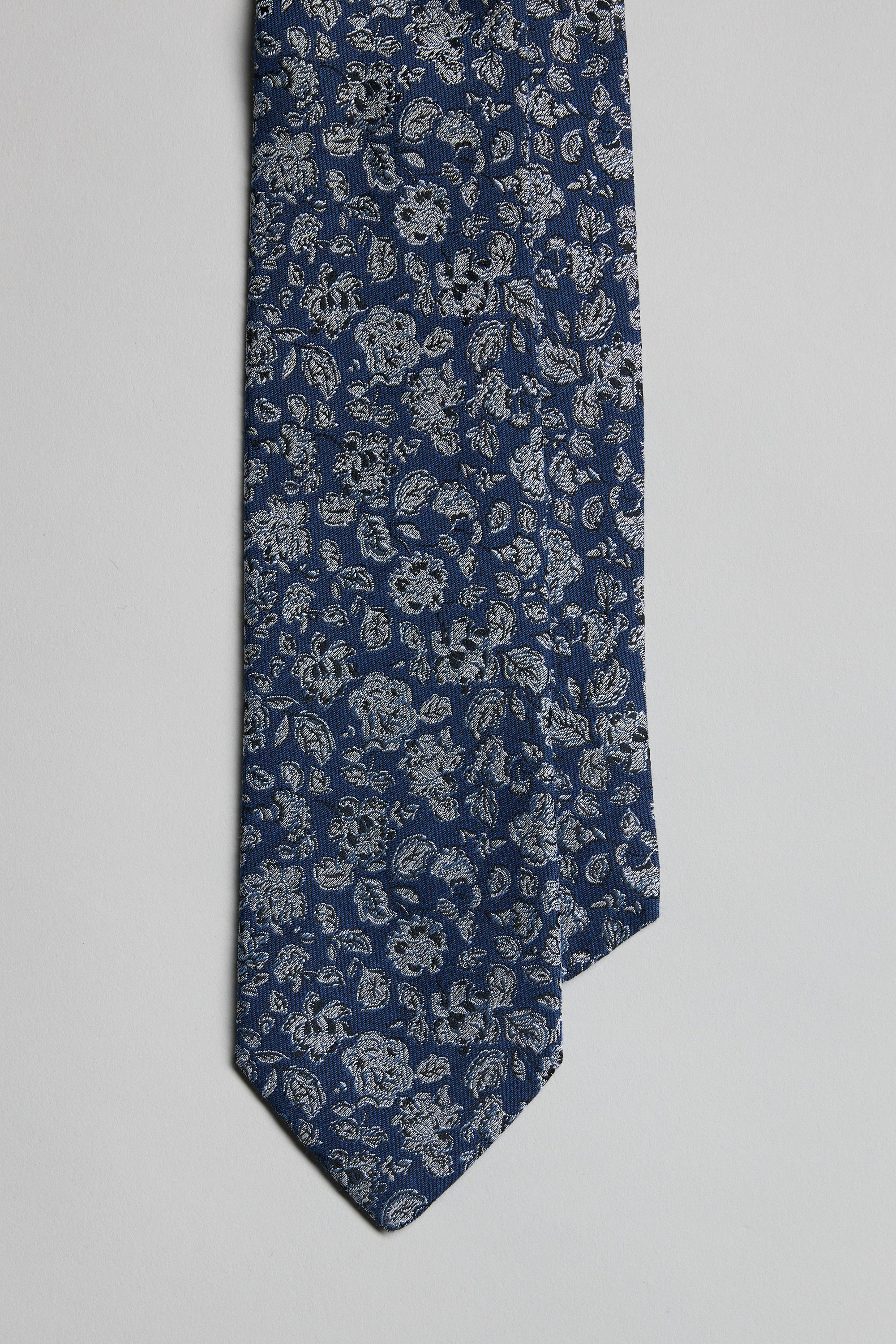 The Parisian Floral Tie by Parisian features a detailed light gray pattern on blue, handmade in New Zealand. Displayed on a plain background, the tie is shown from tip upward with its tip perfectly centered.