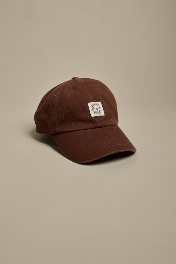 The Barkers Washed Cotton Cap by Barkers is a brown adjustable hat featuring a small white square patch with a circular logo on the front, shown on a plain beige background. It offers comfort and style for everyday wear.