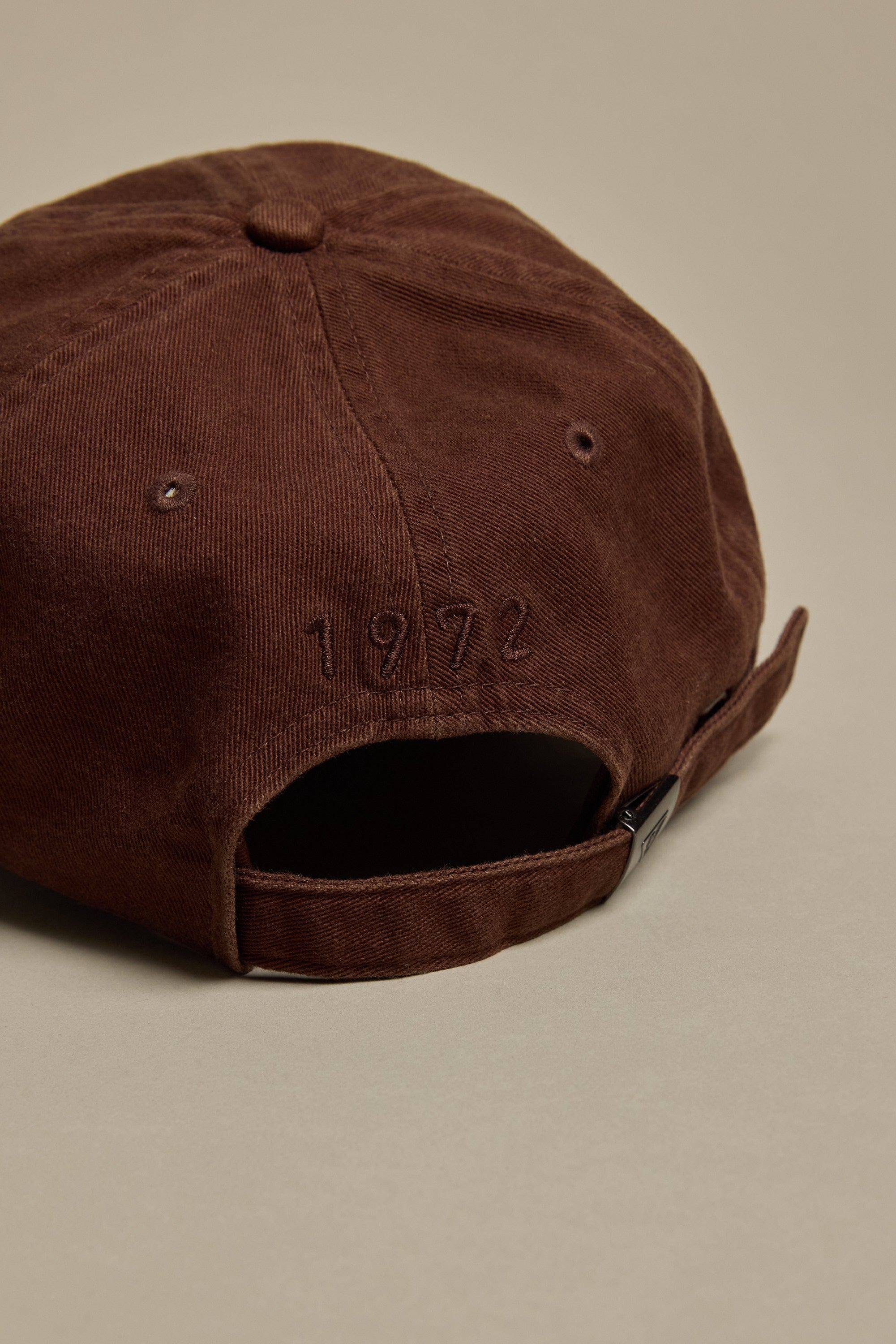 The Barkers Washed Cotton Cap by Barkers is shown from the back in brown, featuring '1976' embroidered above an adjustable strap with a metal clasp. The background is a plain, neutral surface.