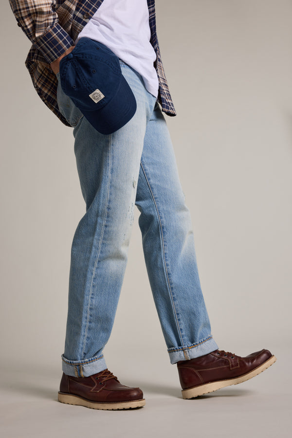 A person in light blue jeans, a white shirt, and a plaid shirt holds the Barkers Washed Cotton Cap by Barkers and wears brown leather boots. The adjustable cap adds style to the neutral studio setting shown from the waist down.