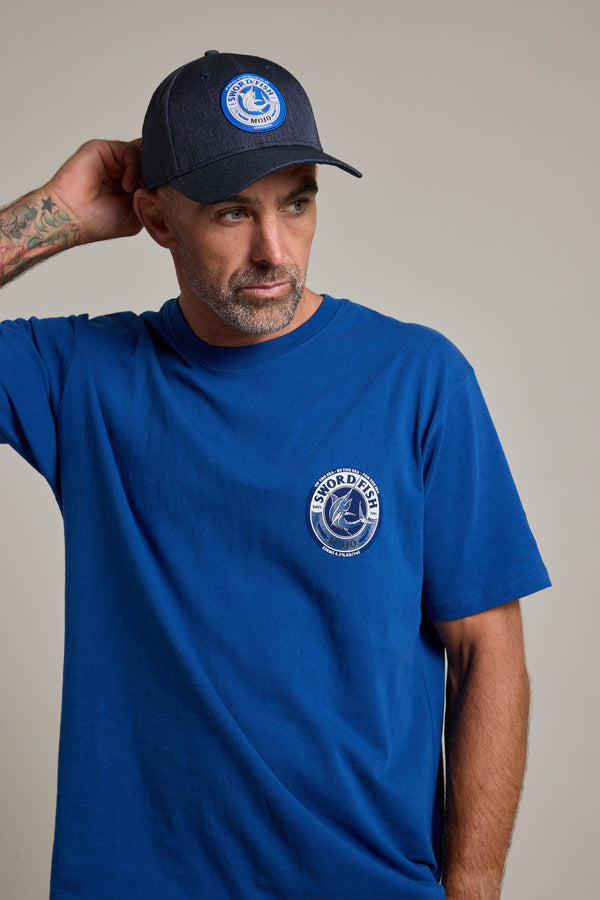 A man in a blue t-shirt and the Swordfish Mojo Trucker Cap by Swordfish Mojo, both displaying the brand's logo, stands against a plain background, looking to the side with one hand behind his head.