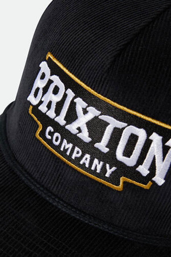 Close-up of the Brixton Pierson Snapback, a black cotton corduroy hat by Brixton, featuring an adjustable snapback and an embroidered BRIXTON COMPANY patch with bold white letters outlined in yellow.