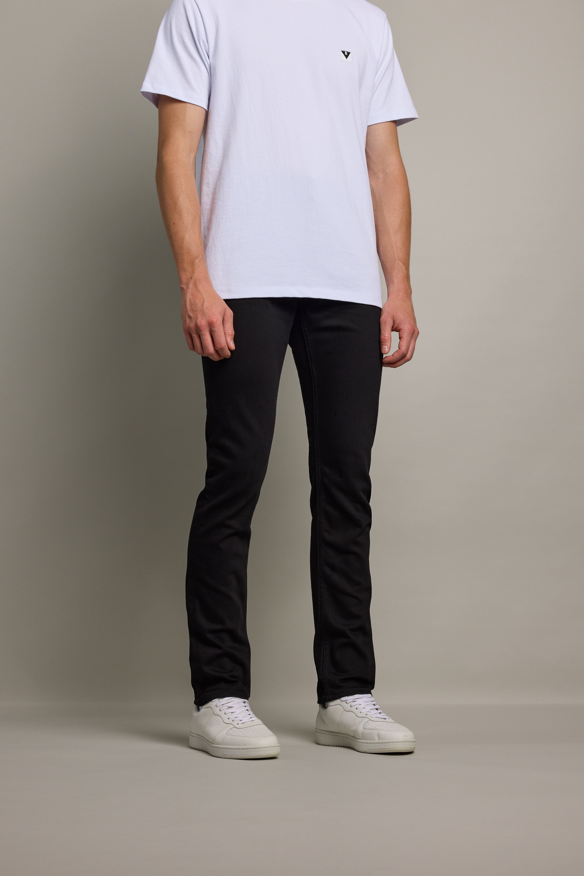 A person stands against a plain background, wearing a white short-sleeve T-shirt, Barkers Rambler Denim Slim Jean in black sustainable denim, and white sneakers. The person's head is not visible in the image.