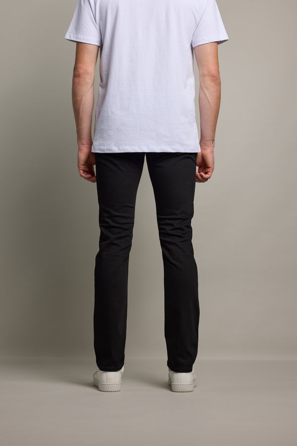 A person stands facing away, wearing a white short-sleeve t-shirt, Barkers Rambler Denim Slim Jean, and white sneakers against a plain, light-colored background.