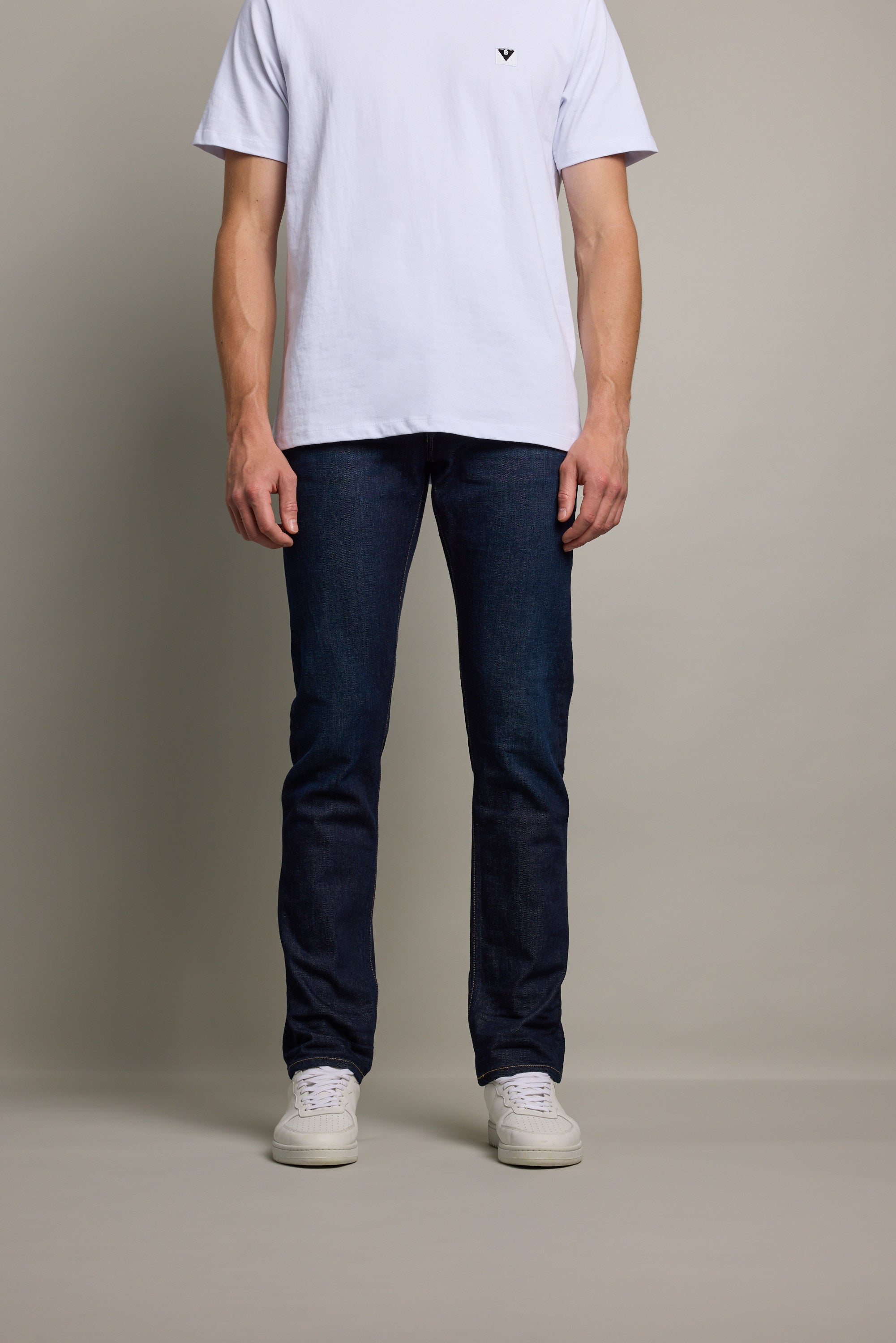 A person wearing a plain white T-shirt, Barkers Rambler Denim Slim Jean, and white sneakers stands against a beige background, with only the lower half of their face to their feet visible.