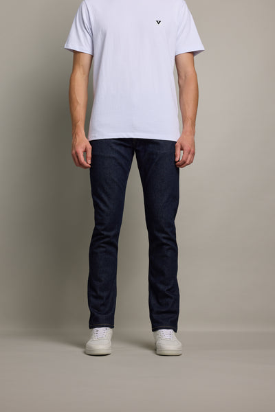 A person stands against a plain background, wearing Barkers Rambler Denim Slim Jean in dark blue, a white short-sleeve t-shirt, and white sneakers. The image is cropped at the shoulders, so the face is not visible.