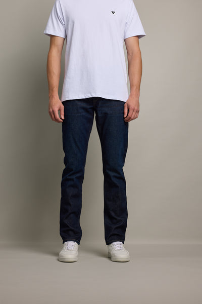 A person stands against a plain background, wearing a white short-sleeve t-shirt, Barkers Nomad Denim Taper Jean in dark blue with an athletic fit, and white sneakers. Their face is not visible.