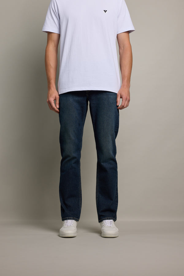 A person stands against a light gray background wearing a plain white T-shirt, Barkers Wanderer Denim Straight Jean in sustainable denim, and white sneakers. The image is cropped at the shoulders.