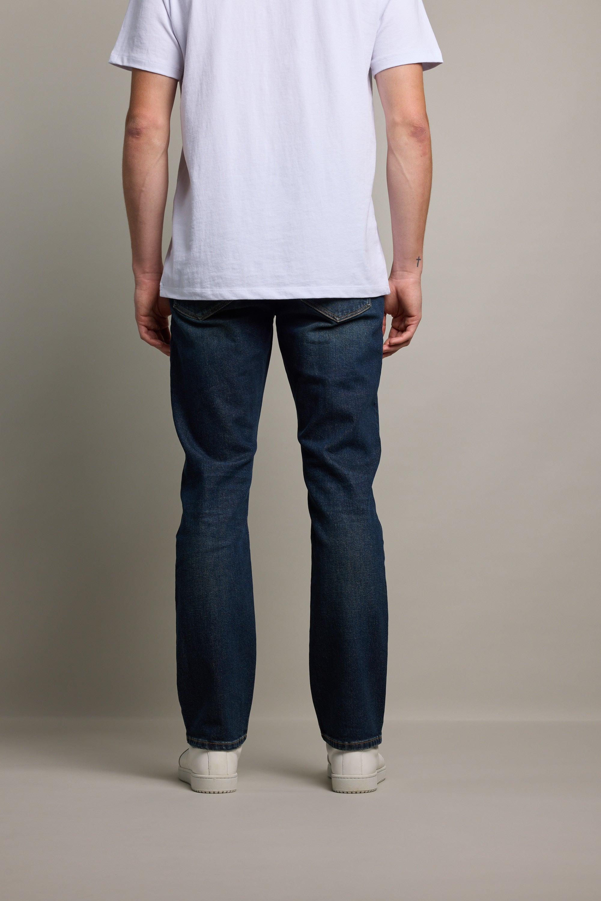 A person stands facing away from the camera, wearing a white t-shirt, Barkers Wanderer Denim Straight Jean made from sustainable denim, and white shoes, against a plain light background.