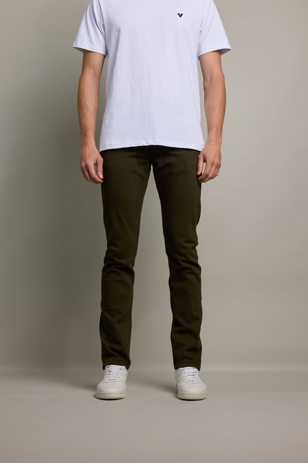 A person stands against a plain background wearing a white t-shirt, Barkers Rambler Slim Coloured Jean made with regenerative cotton from Saitex Vietnam, and white sneakers. Their head is not visible in the image.