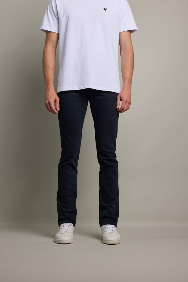 A person stands against a plain background wearing a white t-shirt, Barkers Rambler Slim Coloured Jean made from regenerative cotton by Saitex Vietnam, and white sneakers. The image shows the person from shoulders to feet.