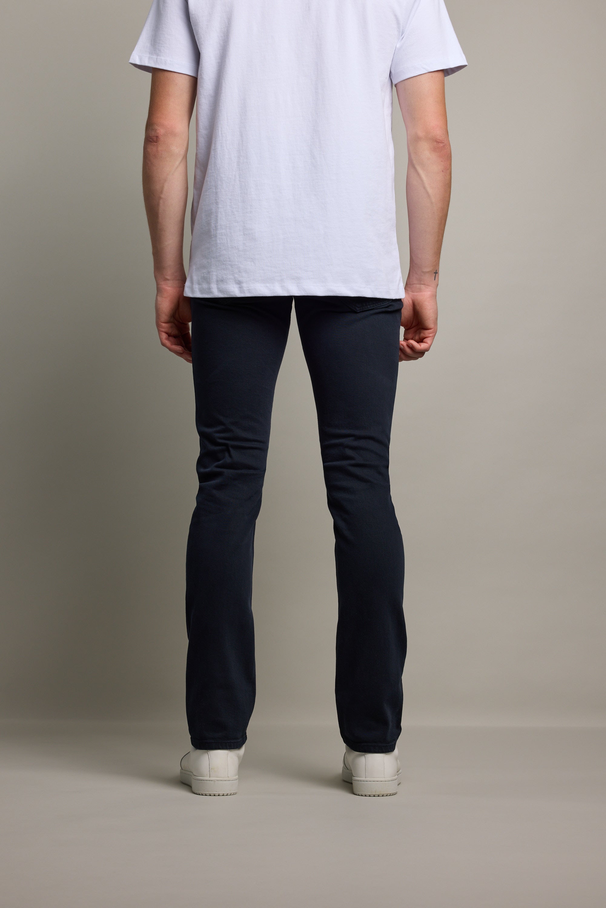 A person stands facing away from the camera, wearing a white T-shirt, Barkers Rambler Slim Coloured Jean made with regenerative cotton by Saitex Vietnam, and white sneakers, against a plain light background.