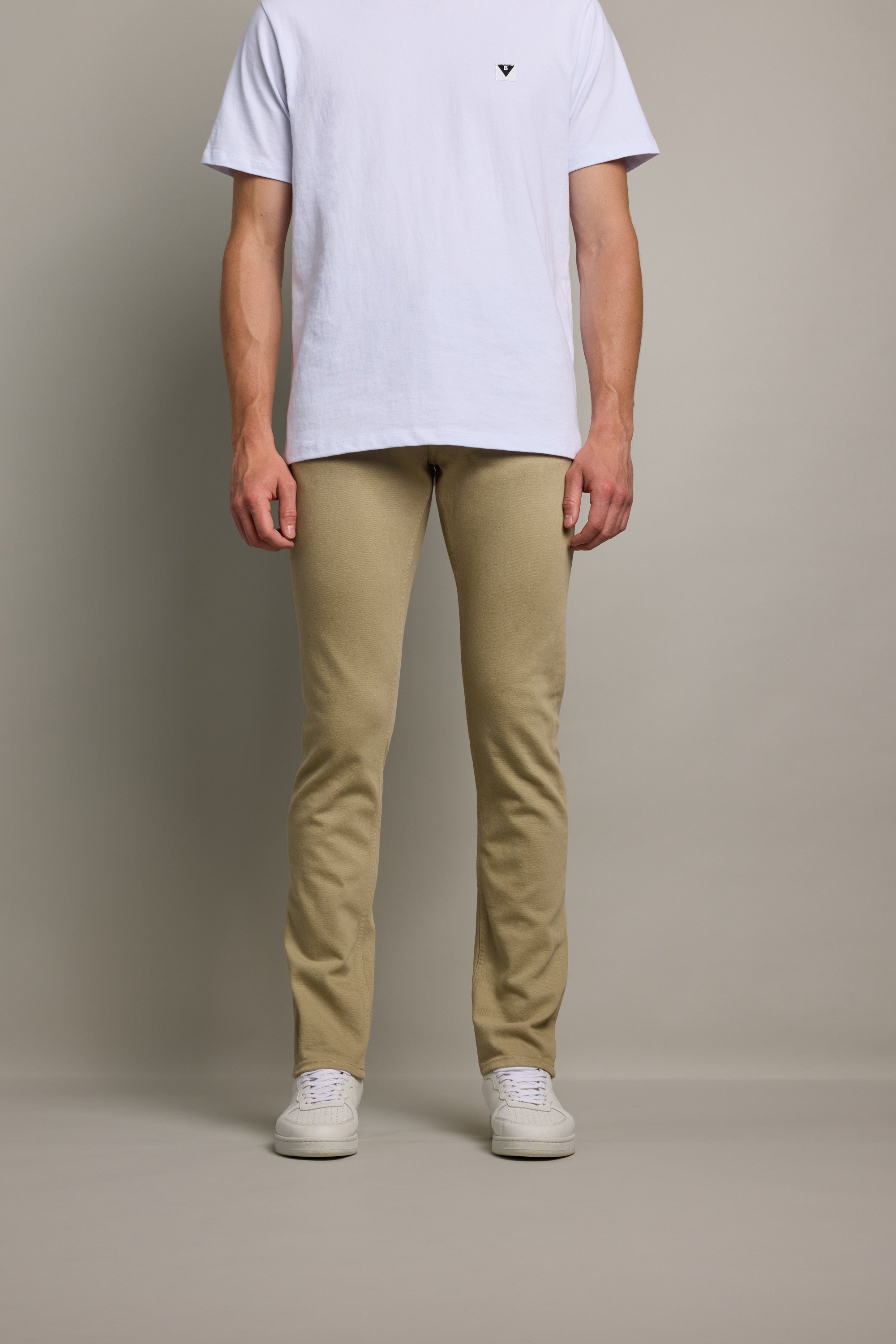 A person stands against a plain background in a white T-shirt, beige pants, and white sneakers. For a modern, eco-conscious look, pair with Barkers' Rambler Slim Coloured Jean. The image is cropped at the shoulders.