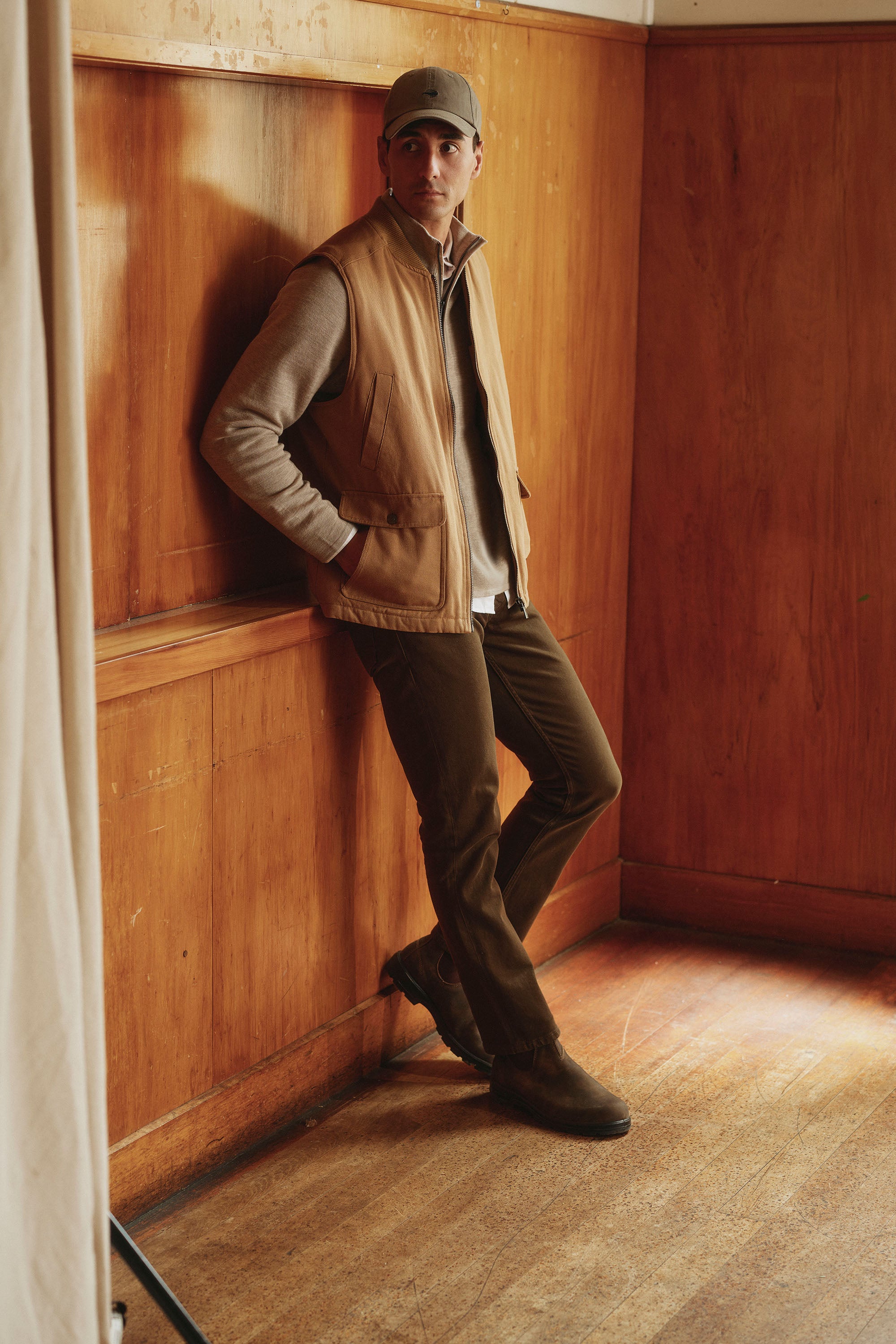 A man in a gray cap, tan vest, light gray sweater, and Barkers Wanderer Straight Coloured Jean made from regenerative cotton leans casually against a wood-paneled wall in a warmly lit room with wooden floors.