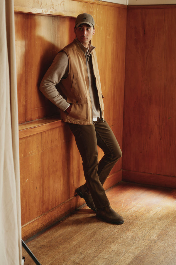 A man in a gray cap, tan vest, light gray sweater, and Barkers Wanderer Straight Coloured Jean made from regenerative cotton leans casually against a wood-paneled wall in a warmly lit room with wooden floors.