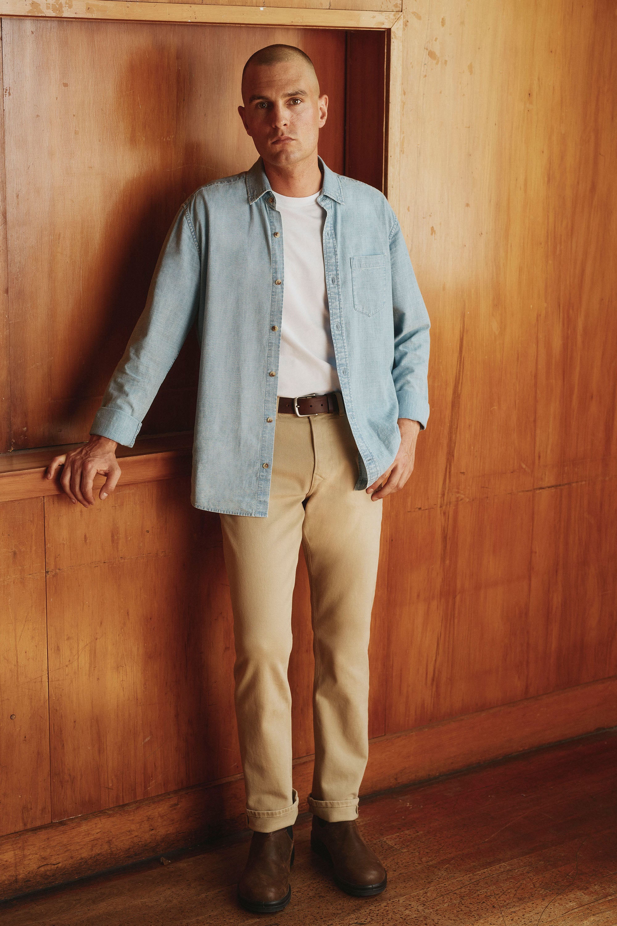 A man with short hair stands against a wooden wall, wearing Barkers’ Wanderer Straight Coloured Jean paired with a light blue sustainable denim shirt, white t-shirt, and brown boots. His look is completed with a neutral expression.