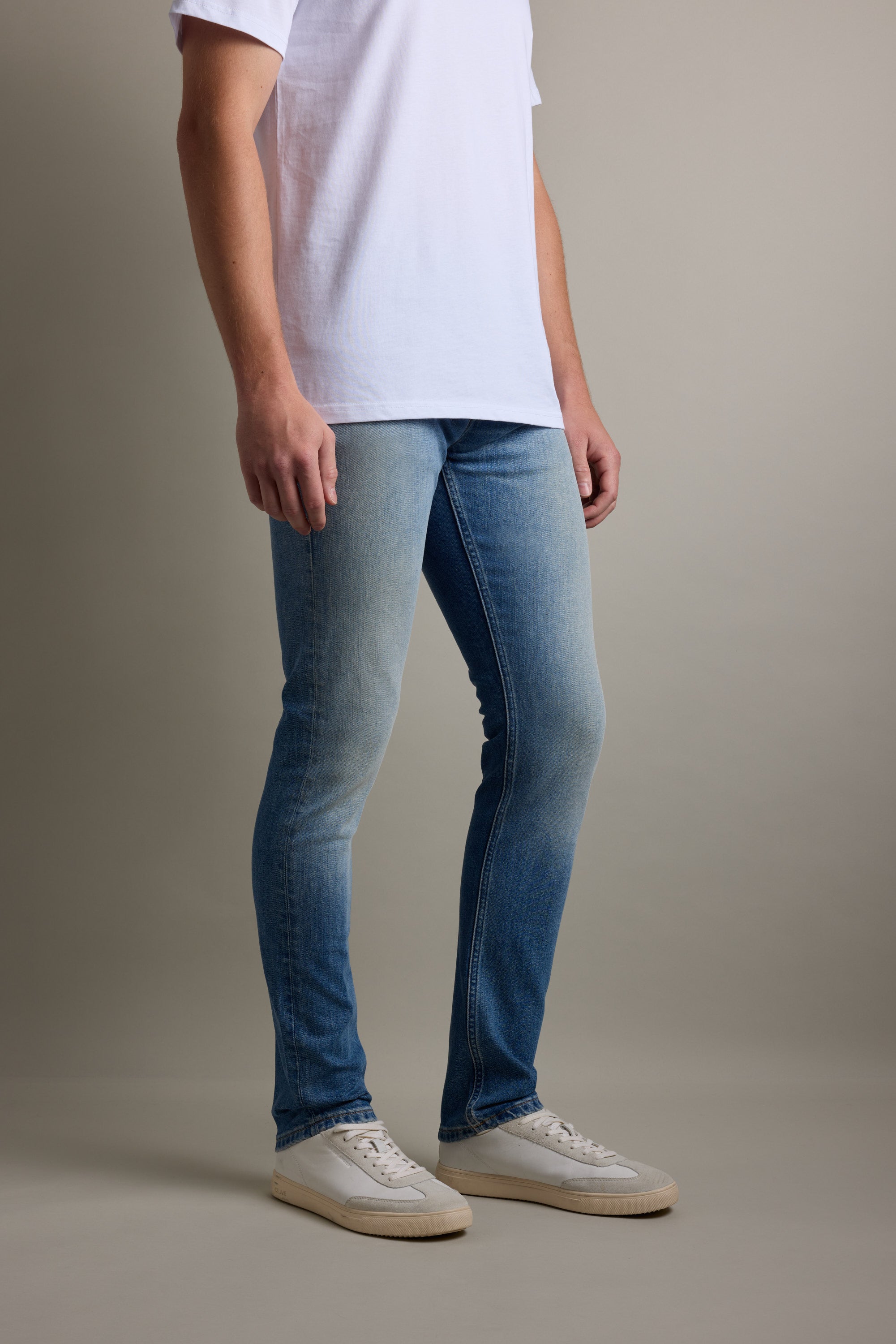 Person wearing Barkers Rambler Denim Slim Jean, white t-shirt, and white sneakers indoors.