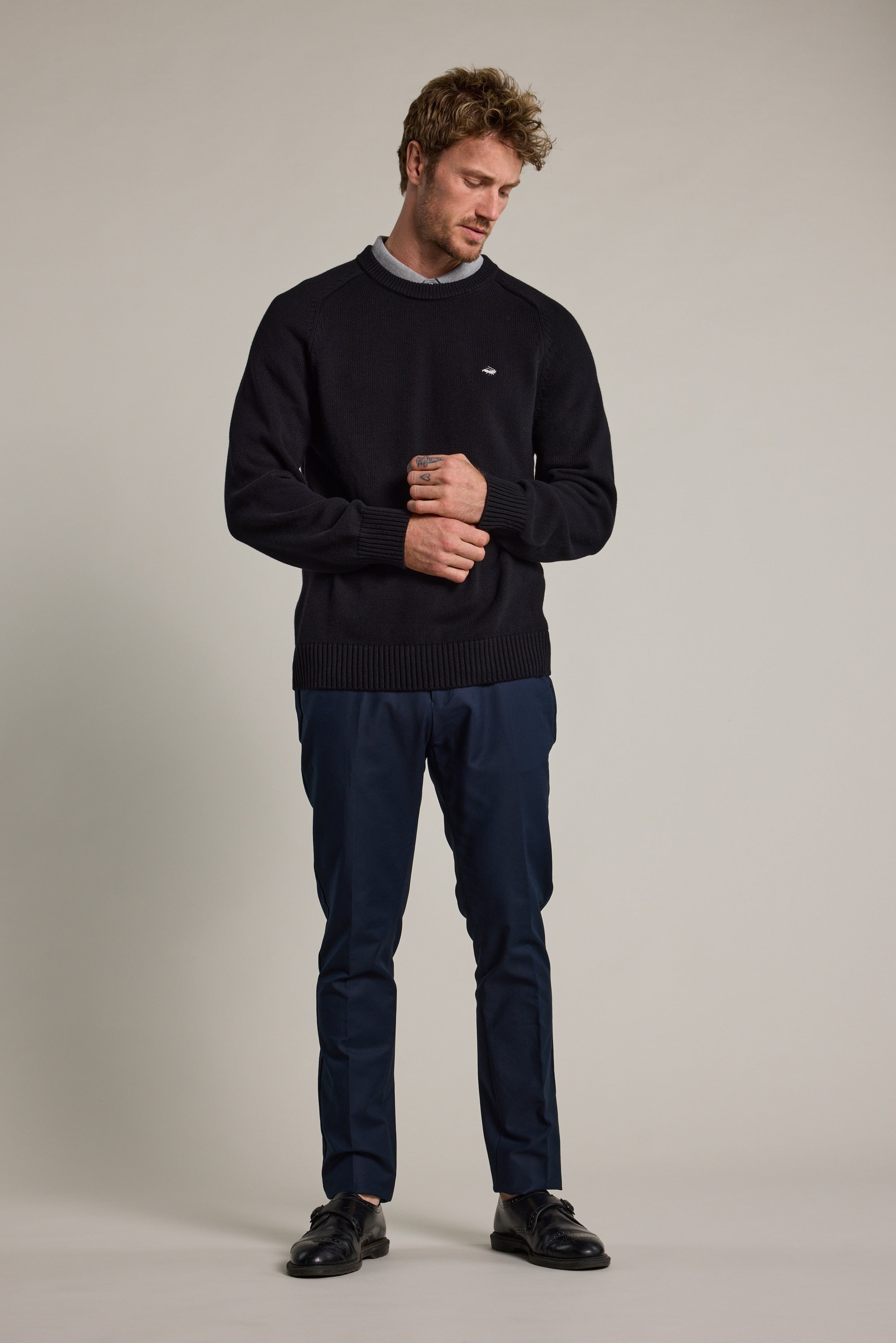 A man stands against a plain background in Barkers Lyndon Cotton Pants, styled with a black sweater over a gray shirt and black shoes. He looks down with a slight smile, his hands loosely clasped in front of him.