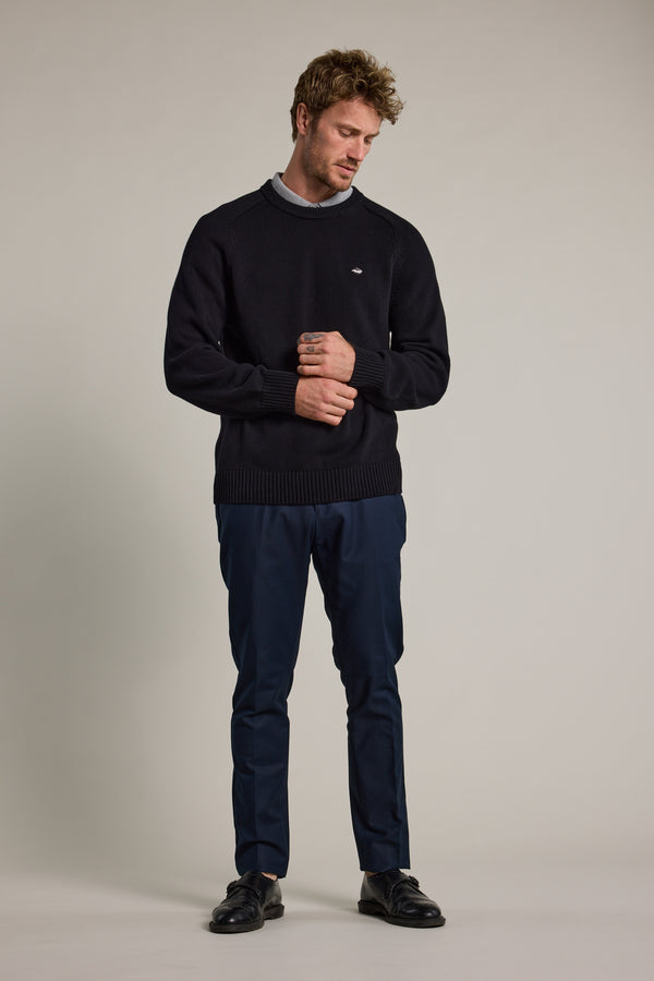 A man stands against a plain background in Barkers Lyndon Cotton Pants, styled with a black sweater over a gray shirt and black shoes. He looks down with a slight smile, his hands loosely clasped in front of him.
