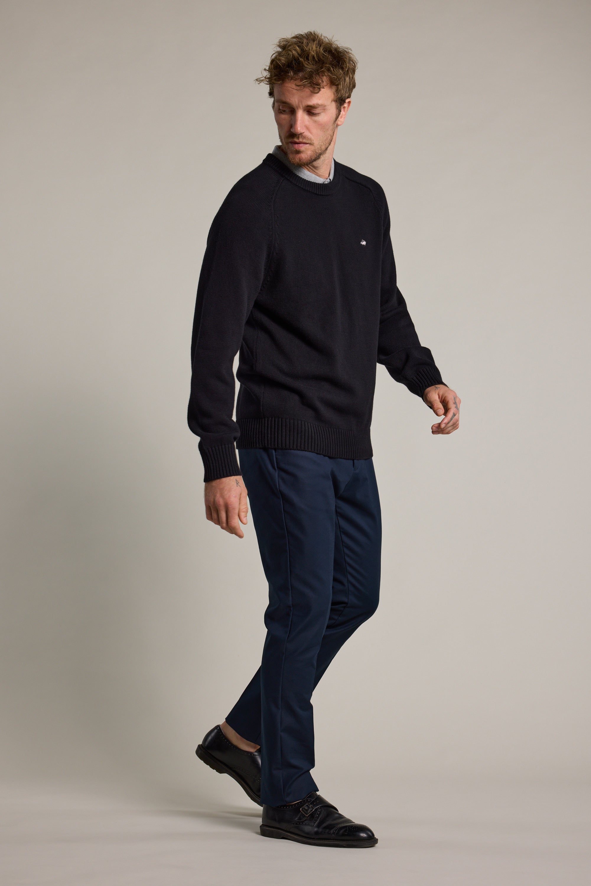 A man with short, tousled hair wears a black sweater, Barkers’ Lyndon Cotton Pants in blue, and black shoes. He stands against a plain light backdrop, looking down to his right for a smart casual look.