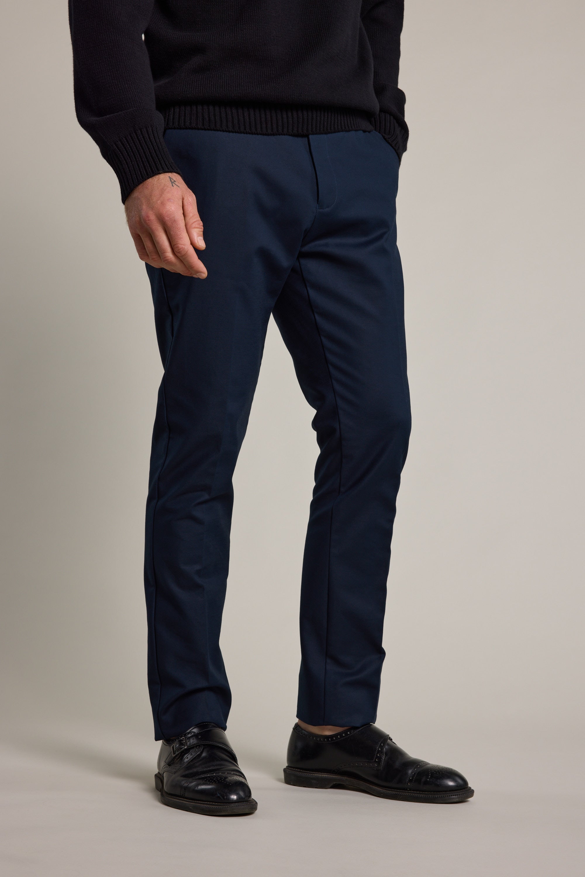 A person stands against a plain light background with hands in pockets, wearing Barkers’ Lyndon Cotton Pants in navy and black dress shoes; only the lower body is visible.