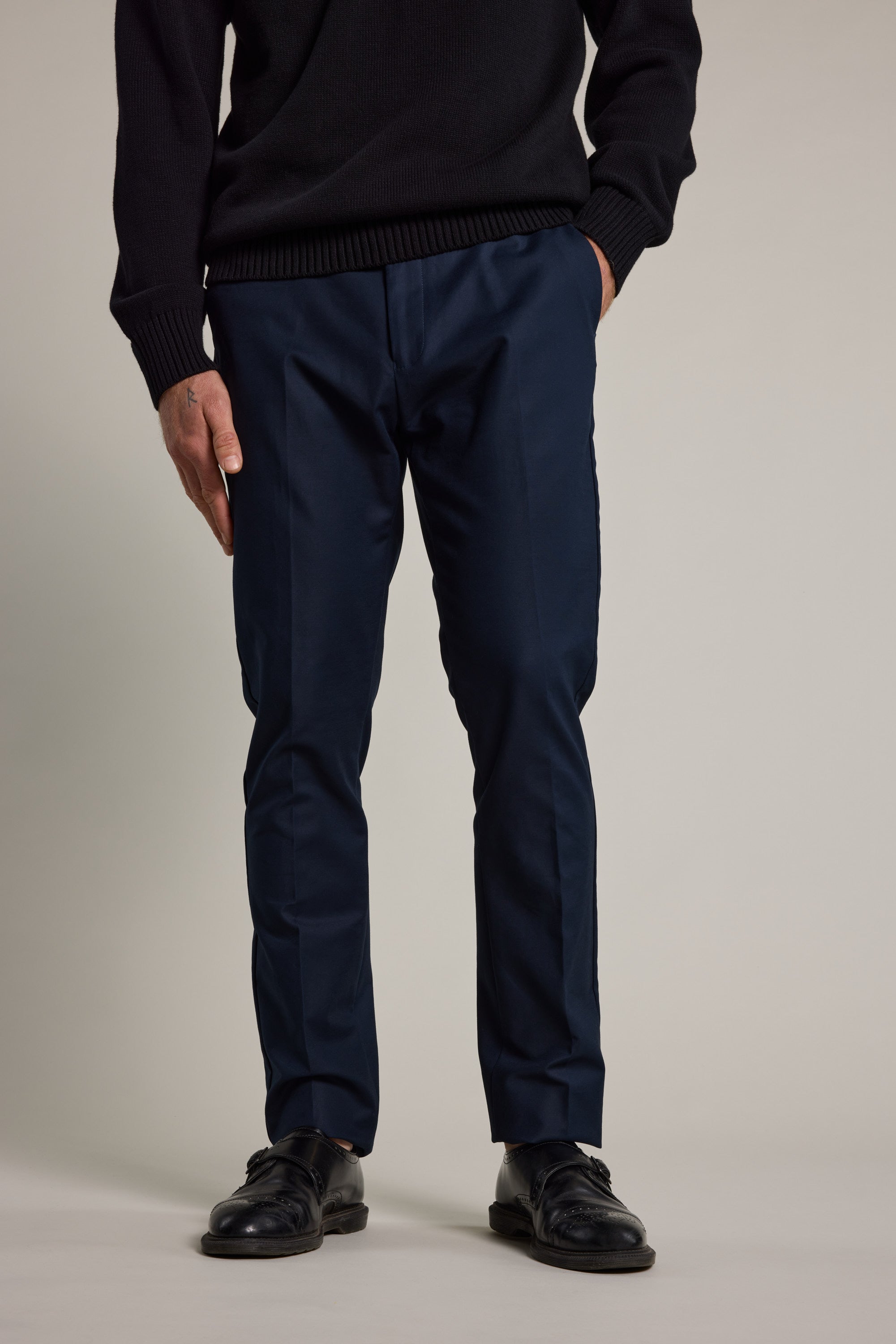 A person wearing a black sweater, Barkers Lyndon Cotton Pants in navy, and black dress shoes stands with hands in pockets against a plain light background. The image is cropped at the shoulders.