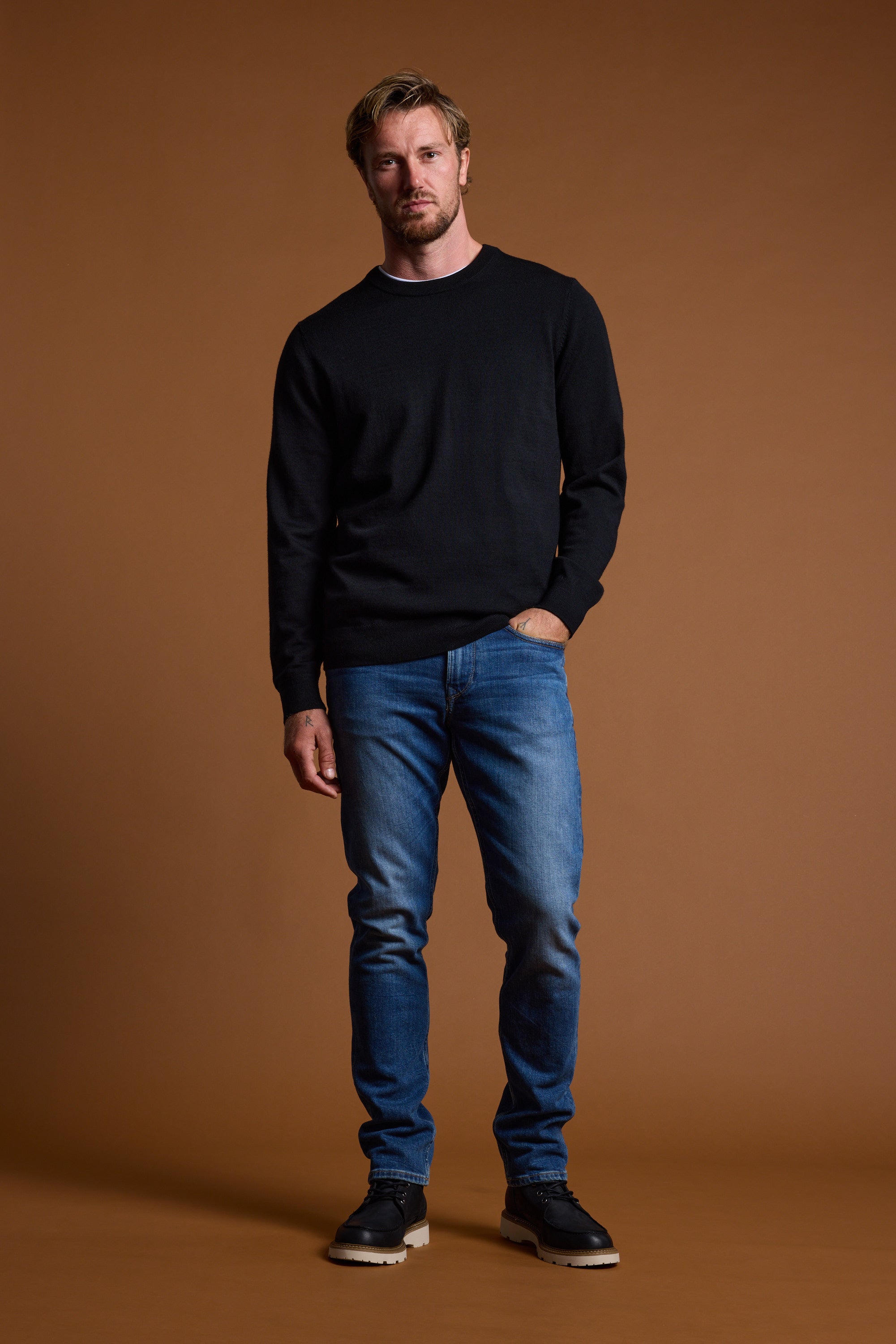 A man with short blond hair and light stubble stands against a brown background wearing the Barkers Skyland ZQrx Merino Crew, blue jeans, and black shoes, one hand in his pocket as he looks at the camera.
