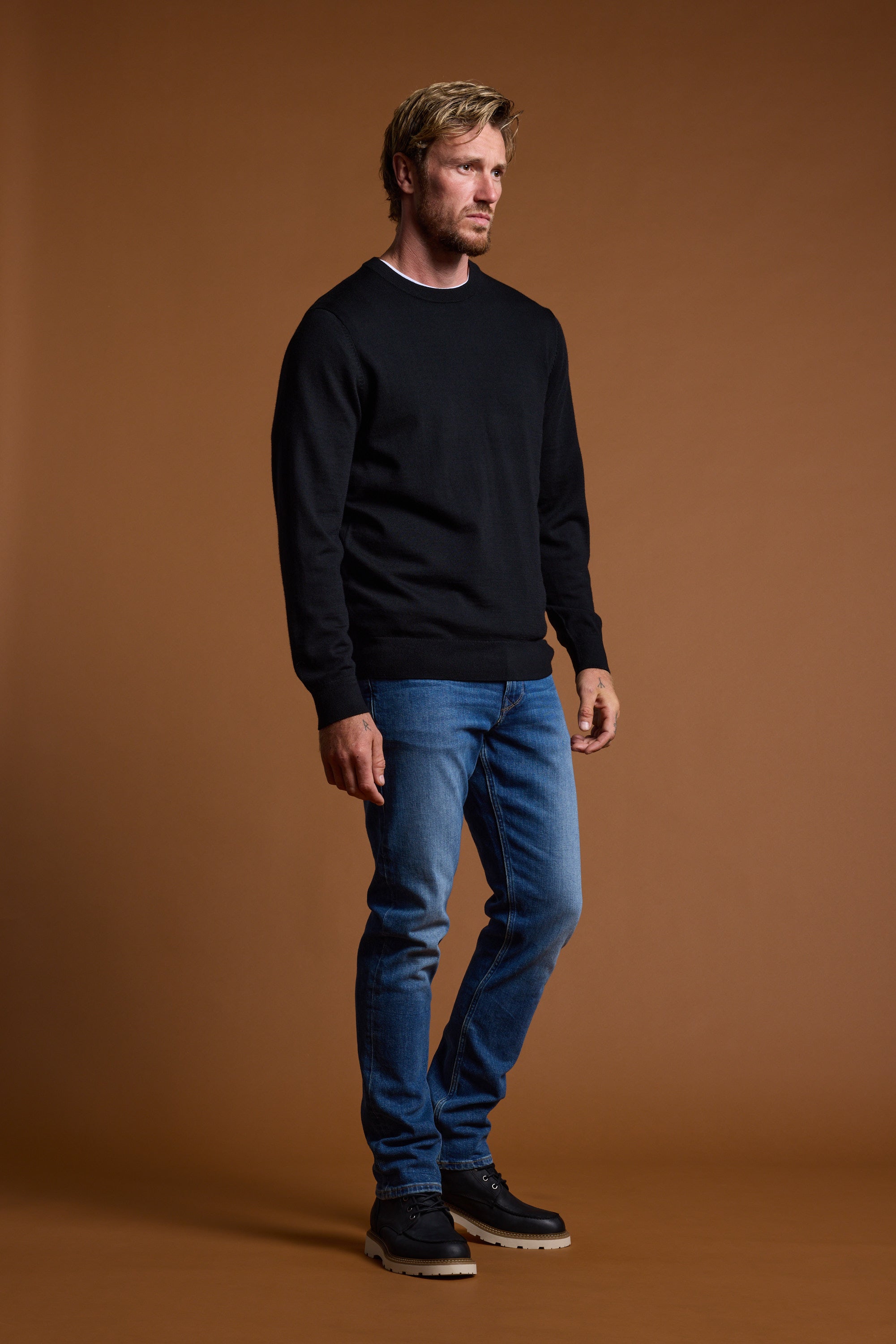 A man with short blond hair and a beard stands against a plain brown background, wearing the Barkers Skyland ZQrx Merino Crew sweater, blue jeans, and black shoes. He gazes off to the side with a neutral expression.