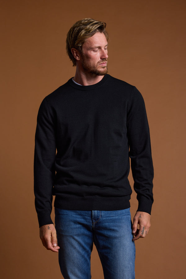 A man with light hair and a beard wears the Barkers Skyland ZQrx Merino Crew in black and blue jeans. He stands against a plain brown background, looking to his left with a neutral expression.
