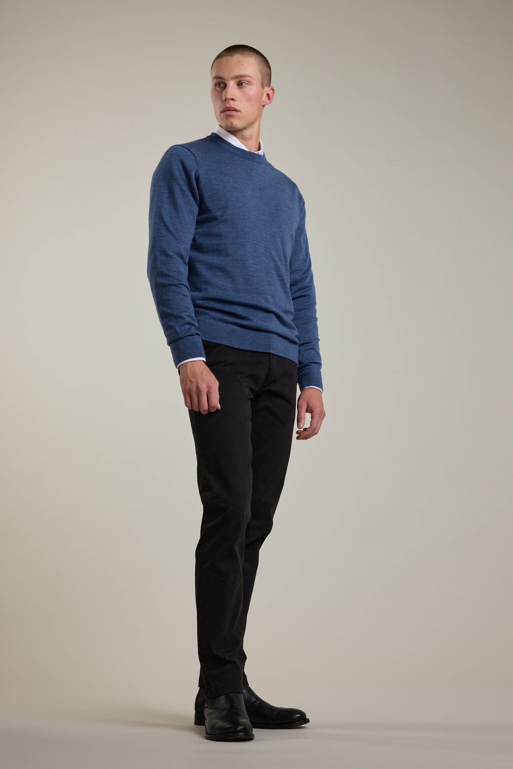 A man stands against a plain light background, wearing a Barkers Skyland ZQrx Merino Crew—crafted from ethically sourced New Zealand merino wool—over a white-collared shirt with black pants and shoes, looking slightly left with a neutral expression.