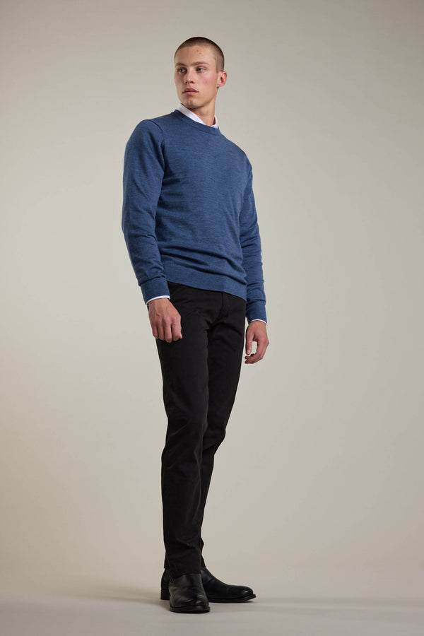 A man stands against a plain light background, wearing a Barkers Skyland ZQrx Merino Crew—crafted from ethically sourced New Zealand merino wool—over a white-collared shirt with black pants and shoes, looking slightly left with a neutral expression.