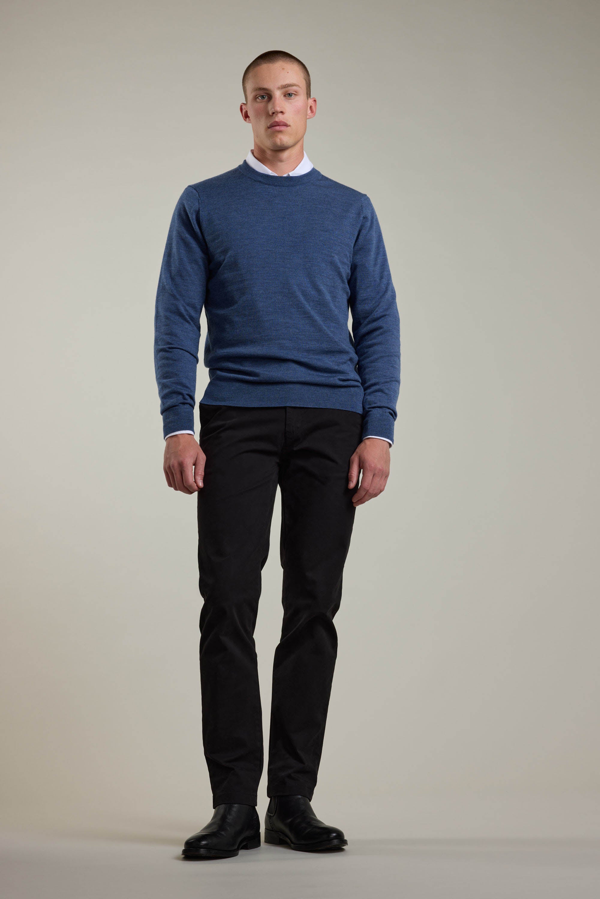 A man faces forward against a plain light background, wearing a Barkers Skyland ZQrx Merino Crew sweater over a white collared shirt, with black pants and shoes. He stands neutrally with his arms by his sides.