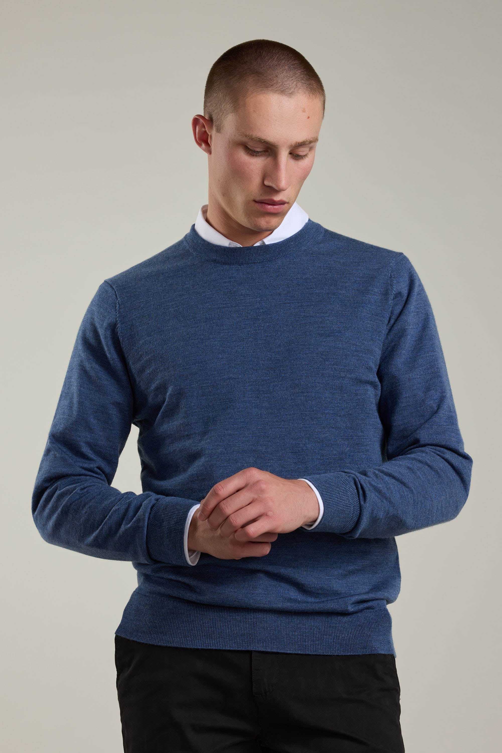 A man with short hair wears a Barkers Skyland ZQrx Merino Crew, made from ethically sourced New Zealand merino wool, over a white collared shirt as he looks down and adjusts his sleeve against a plain light background.