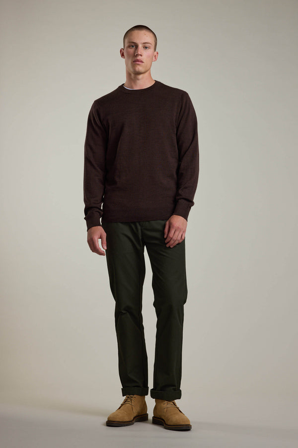 A man with short hair and a neutral expression stands against a plain light background, wearing a dark brown Barkers Skyland ZQrx Merino Crew sweater made from sustainable merino, olive green pants, and tan shoes, arms resting by his sides.
