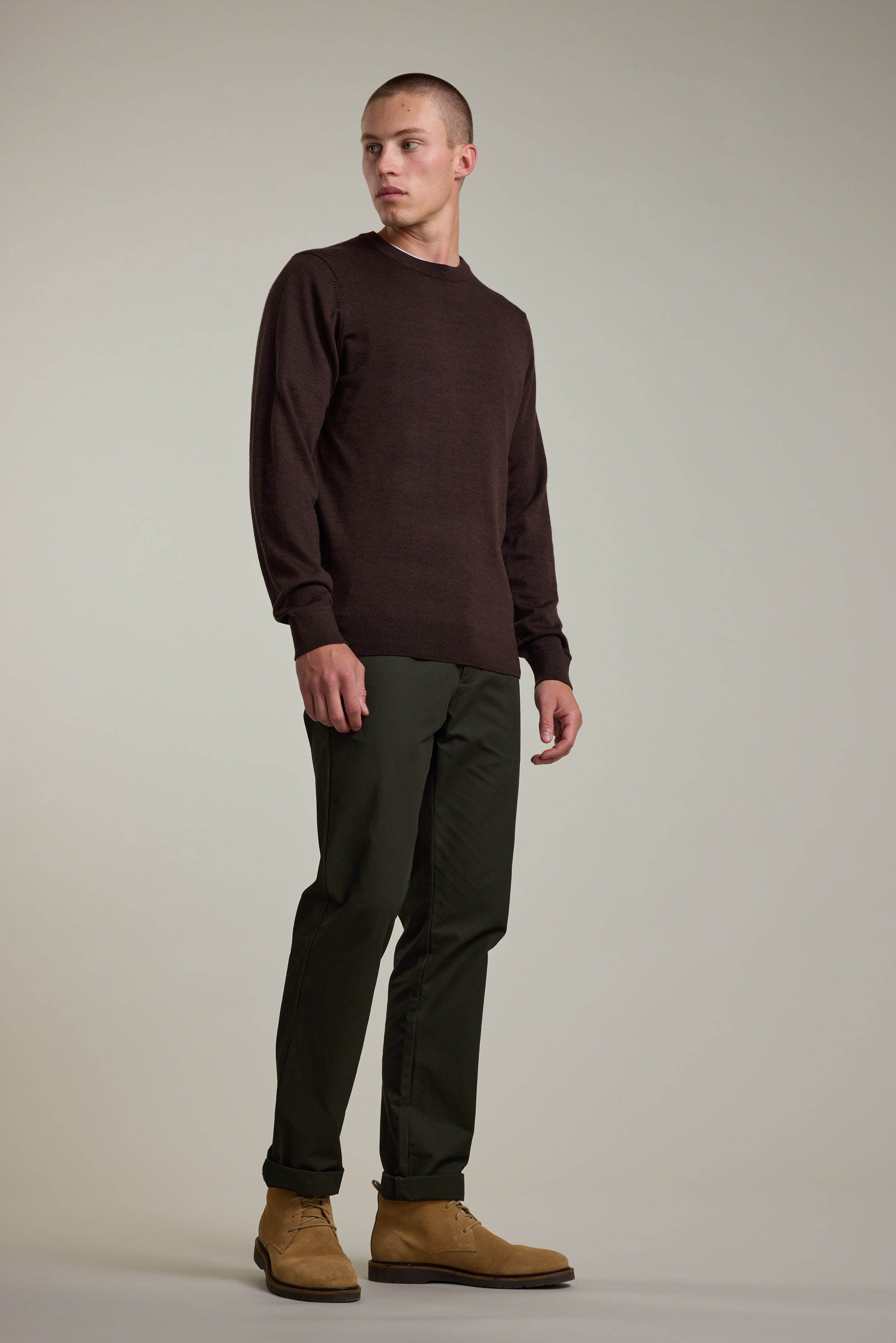 A man stands against a plain light background wearing the Barkers Skyland ZQrx Merino Crew sweater in dark brown, paired with dark green pants and tan suede shoes. He looks slightly to his left with a neutral expression.